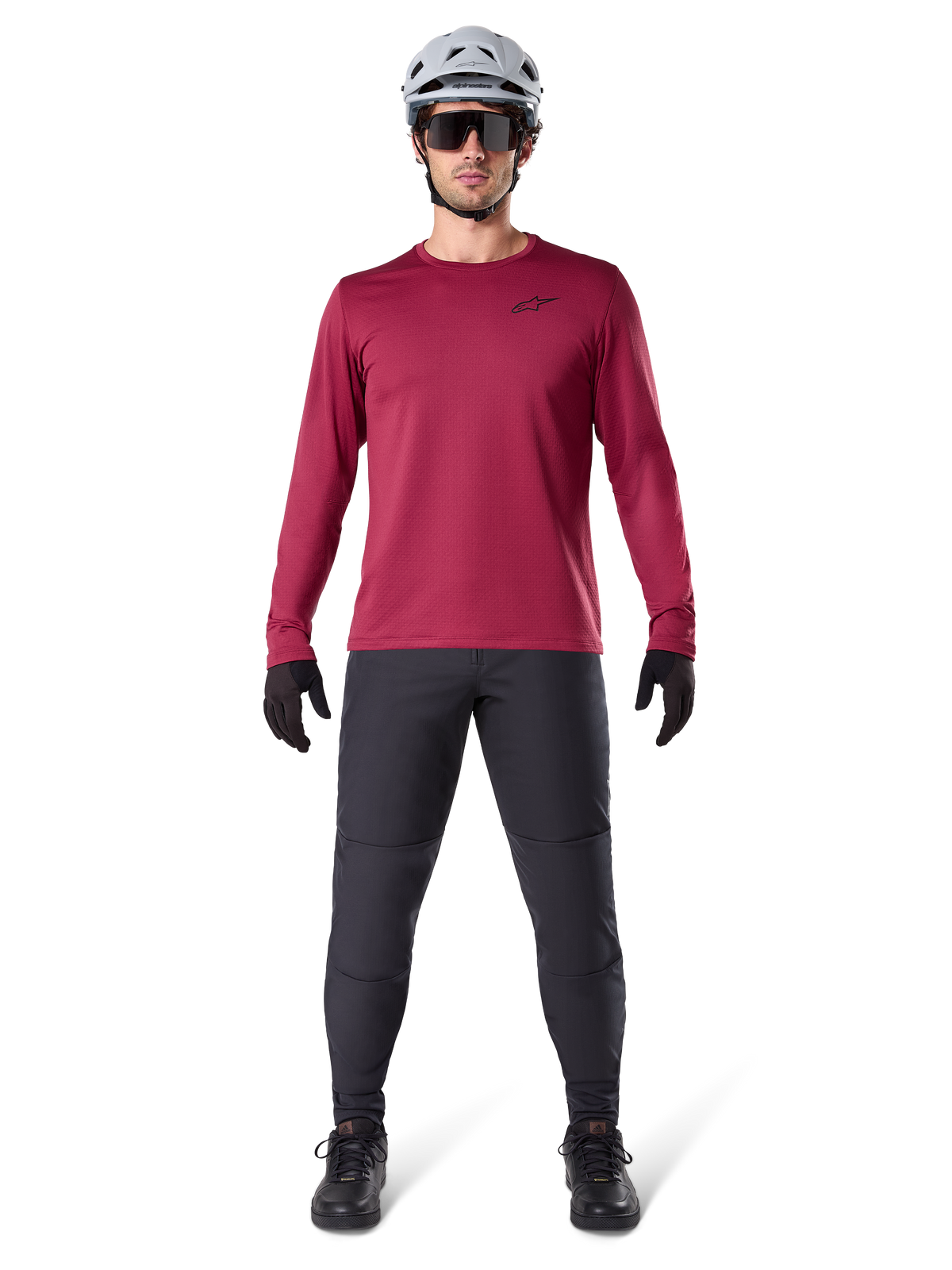 Męski model w koszulce termicznej Alpinestars A-Dura Thermal w kolorze bordowym, koszulka rowerowa z długim rękawem i izolacją, ciemnoszare spodnie MTB, biały kask, czarne okulary przeciwsłoneczne i czarne rękawiczki, profesjonalna odzież MTB na chłodne dni
