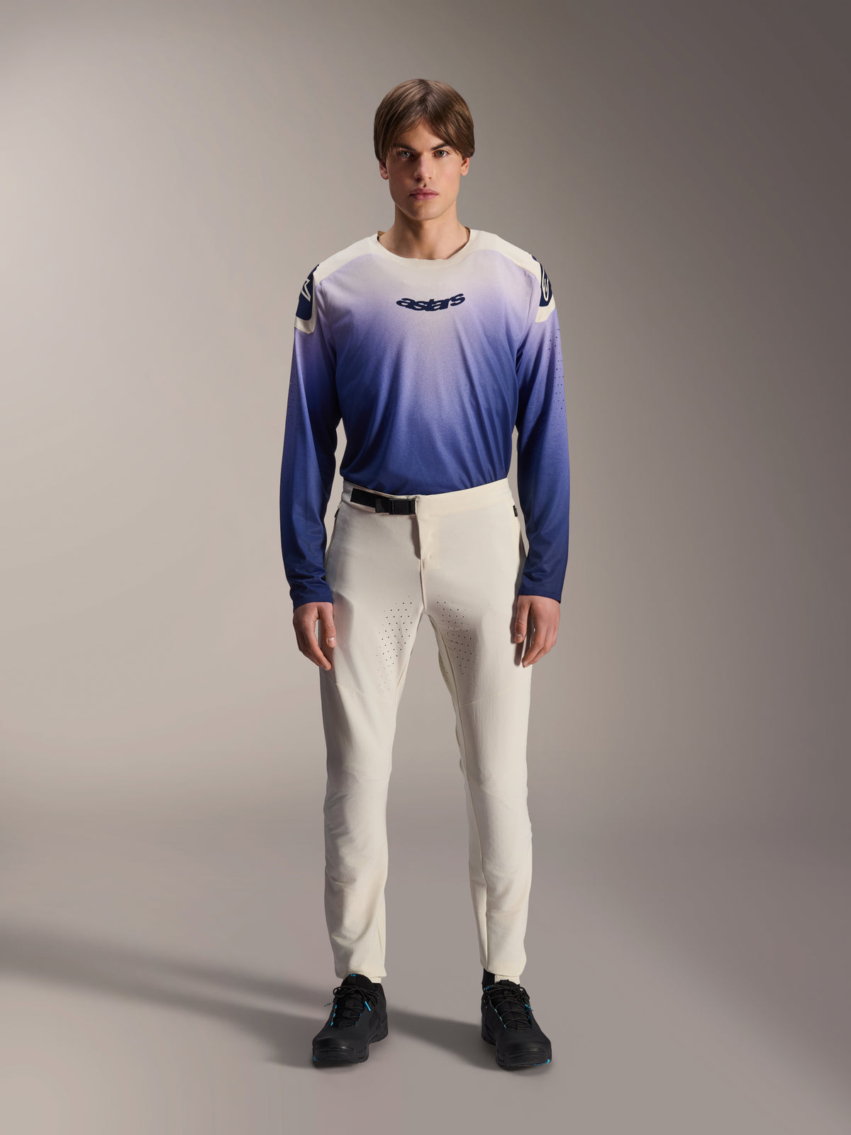 Alpinestars A-Aria Elite Pants, spodnie MTB Performance w kolorze ecru, kremowo-białe spodnie rowerowe z laserowo perforowanymi panelami na udach i magnetyczną klamrą, noszone z niebieską koszulką z długim rękawem z efektem gradientu i czarnymi butami, sportowy krój do jazdy trailowej