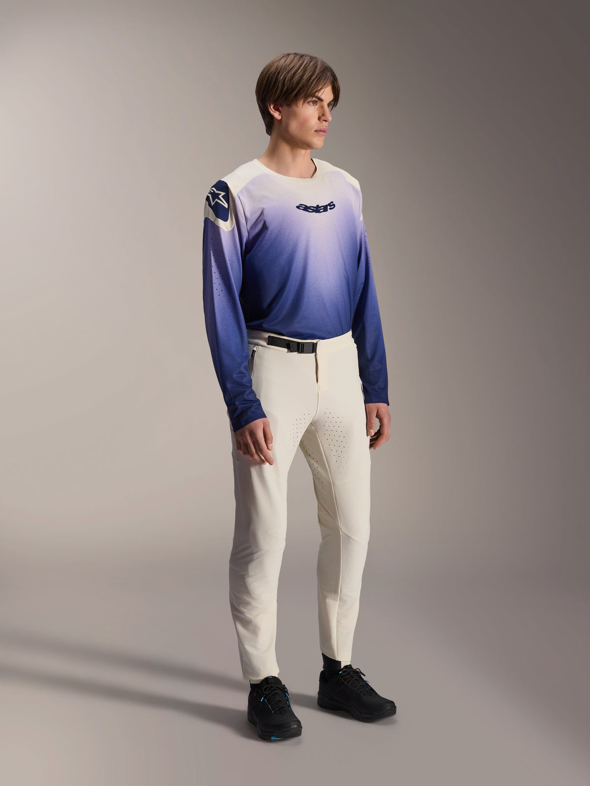 Model ubrany w spodnie Alpinestars A-Aria Elite w kolorze Ecru, kremowe spodnie do kolarstwa górskiego z laserowo wycinaną wentylacją i czarną klamrą w pasie, zestawione z niebiesko-fioletową koszulką z długim rękawem z gradientem, profesjonalna odzież rowerowa na szlaki terenowe