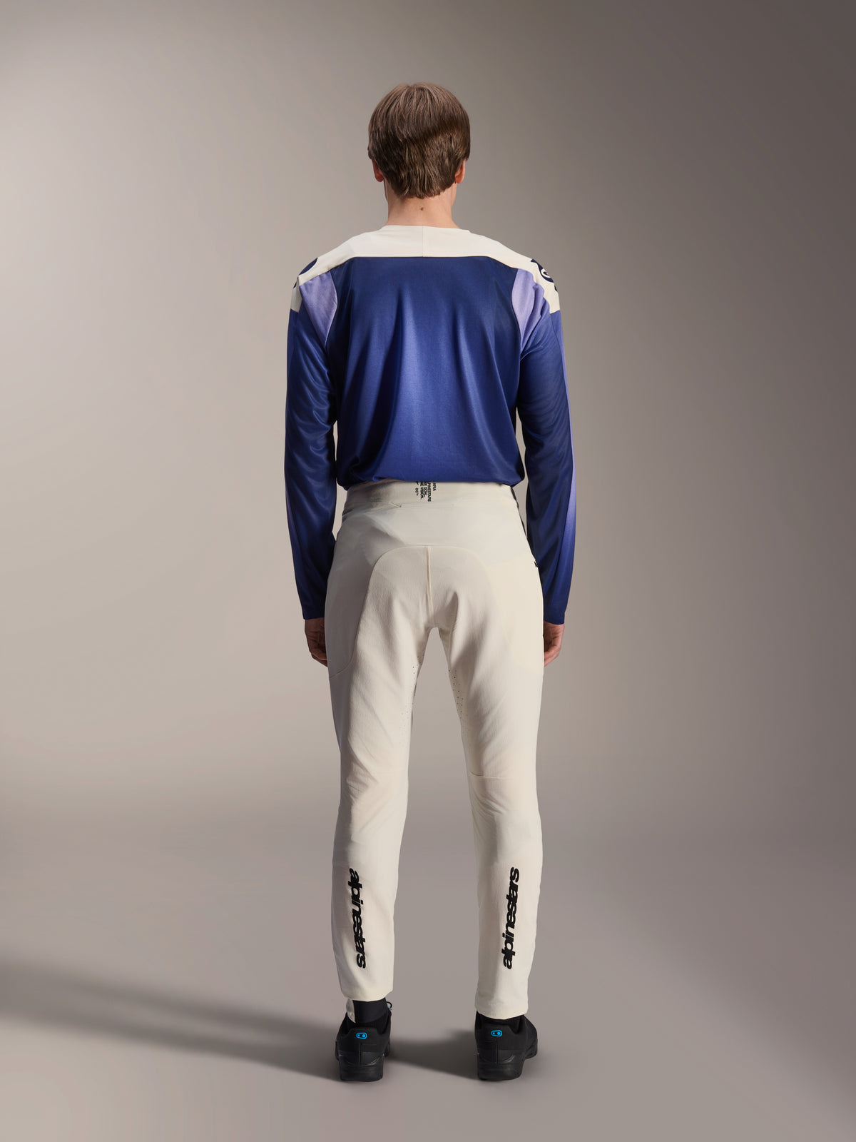 Spodnie Alpinestars A-Aria Elite w kolorze ecru, widok z tyłu na modelu w zestawieniu z niebieską koszulką z długim rękawem A-Aria Polartec Maker Elite, techniczne spodnie MTB w kolorze złamanej bieli, czarne logotypy na dole nogawek, profesjonalna odzież rowerowa do kolarstwa górskiego