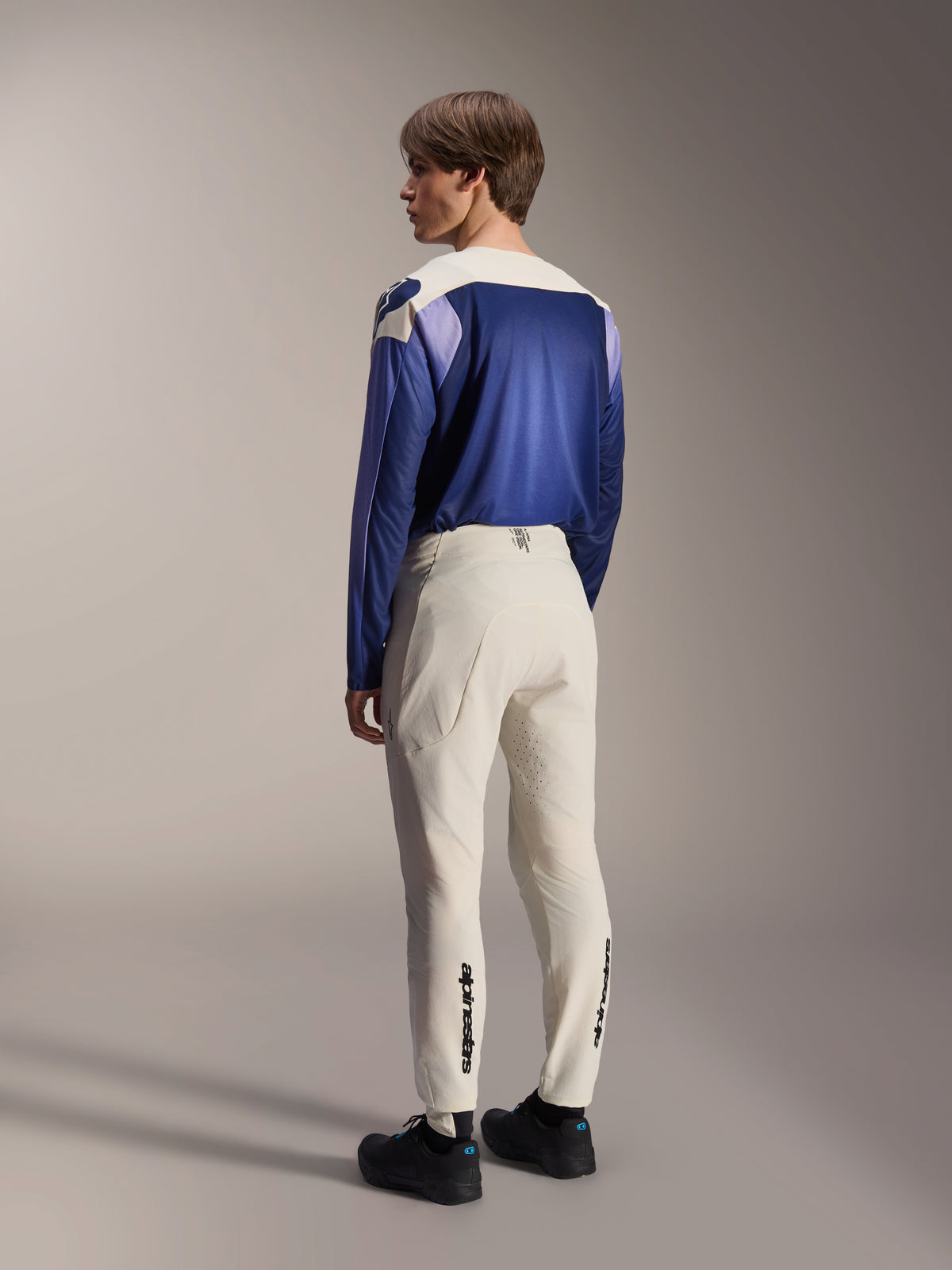 Alpinestars A-Aria Elite Pants, spodnie MTB, kolor ecru, widok z tyłu na modelu, czarny logotyp na dolnej części nogawek i laserowe perforacje dla wentylacji, zestawione z niebiesko-fioletową koszulką z długim rękawem i czarnymi butami, profesjonalna odzież rowerowa