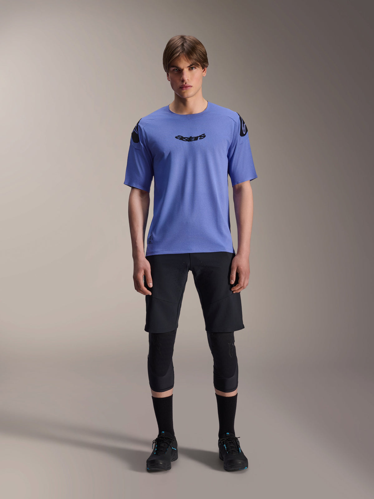 Męski model w koszulce Alpinestars A-Aria Polartec Elite z krótkim rękawem w kolorze Baja Blue, czarne logo astars na klatce piersiowej, czarne spodenki MTB, ochraniacze kolan i czarne buty rowerowe, zdjęcie studyjne odzieży na rower górski