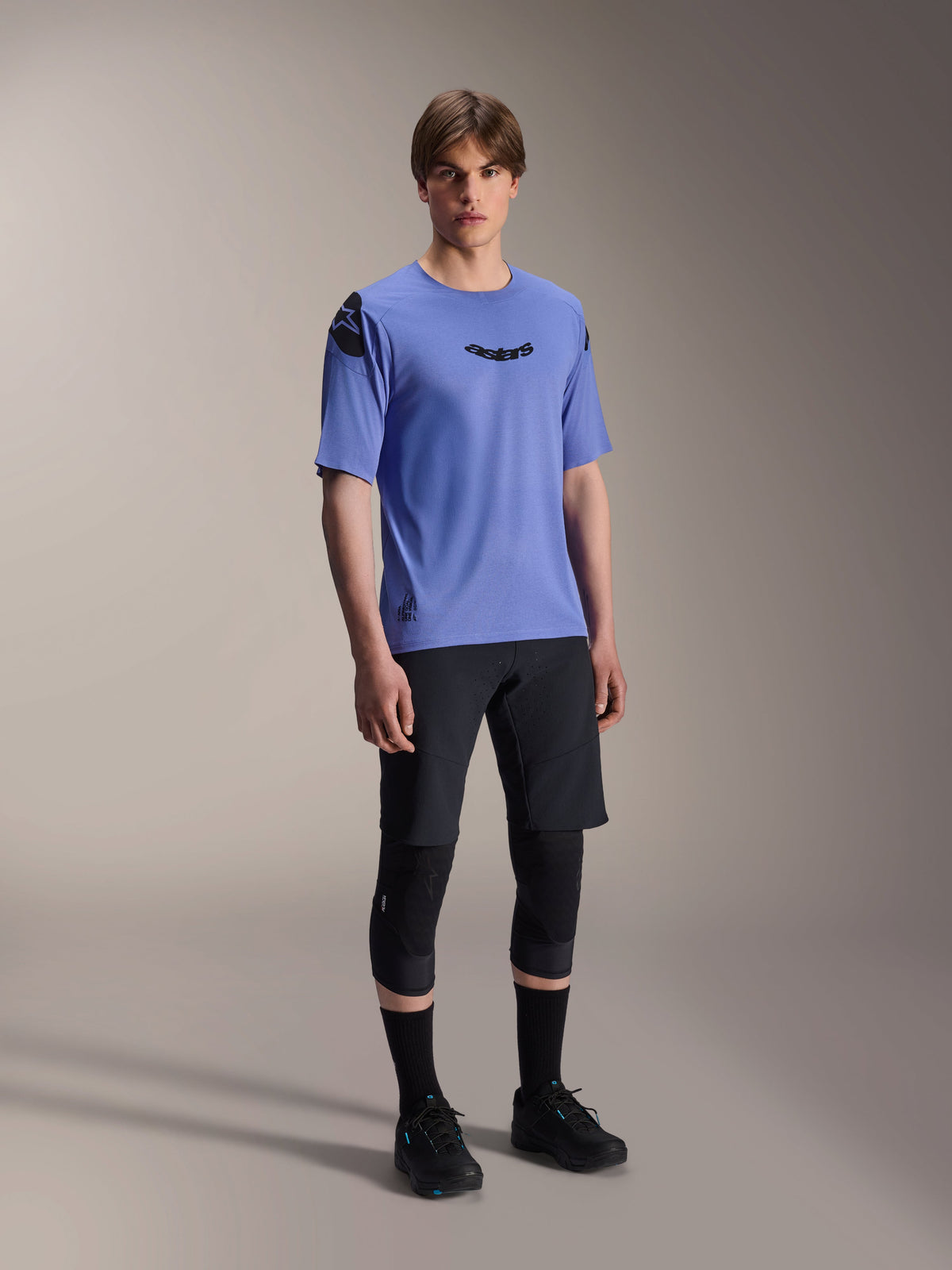 Męski model w koszulce Alpinestars A-Aria Polartec Elite z krótkim rękawem w kolorze Baja Blue, czarne szorty MTB i ochraniacze kolan, czarne logo Astars na klatce piersiowej i ramieniu, profesjonalna odzież rowerowa MTB