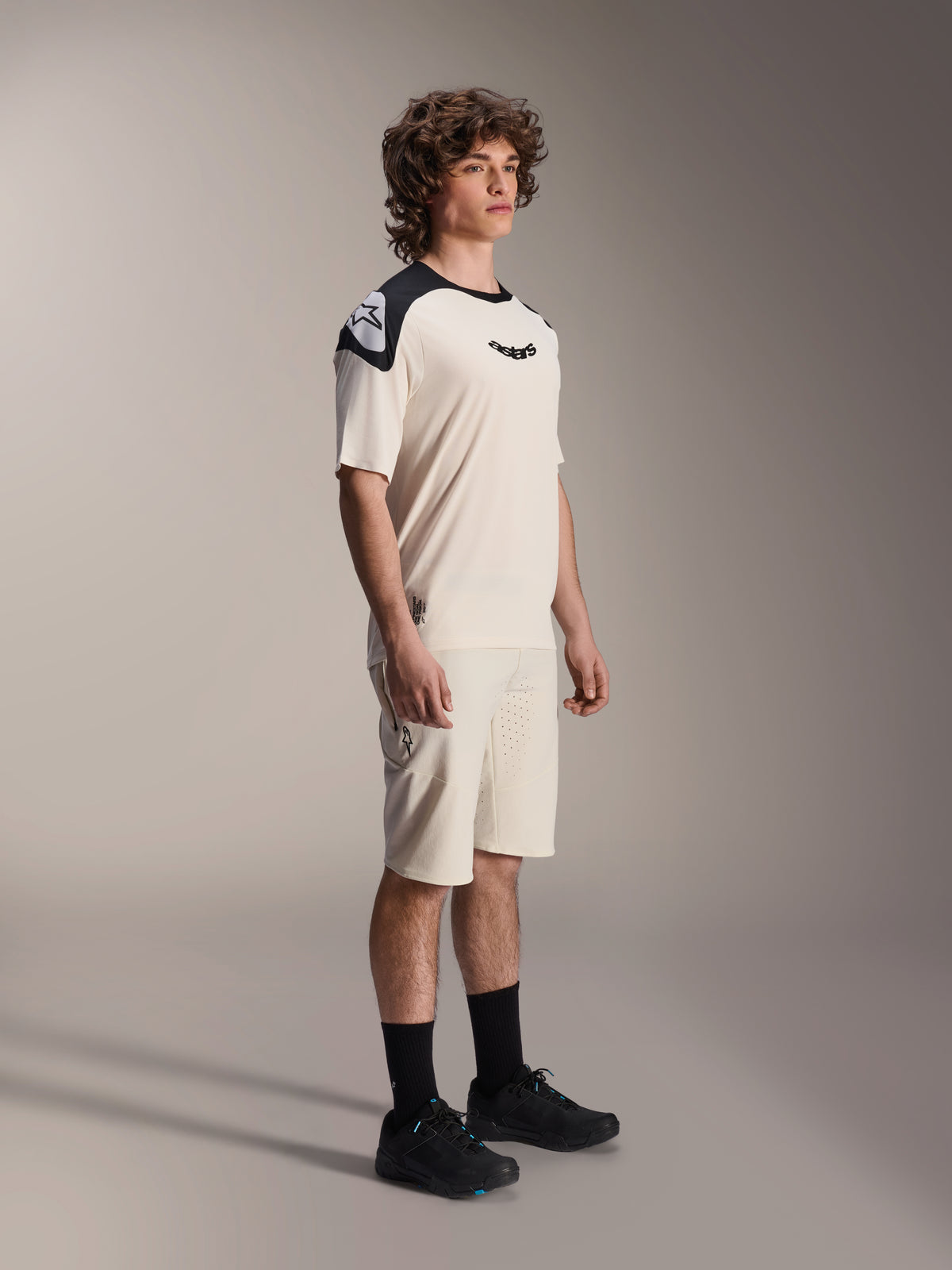 Model męski ubrany w szorty Alpinestars A-Aria Elite w kolorze Ecru, techniczne szorty MTB z laserowo wycinaną wentylacją, zestawione z dopasowaną kremowo-czarną koszulką z krótkim rękawem i czarnymi butami, profesjonalna odzież kolarska.