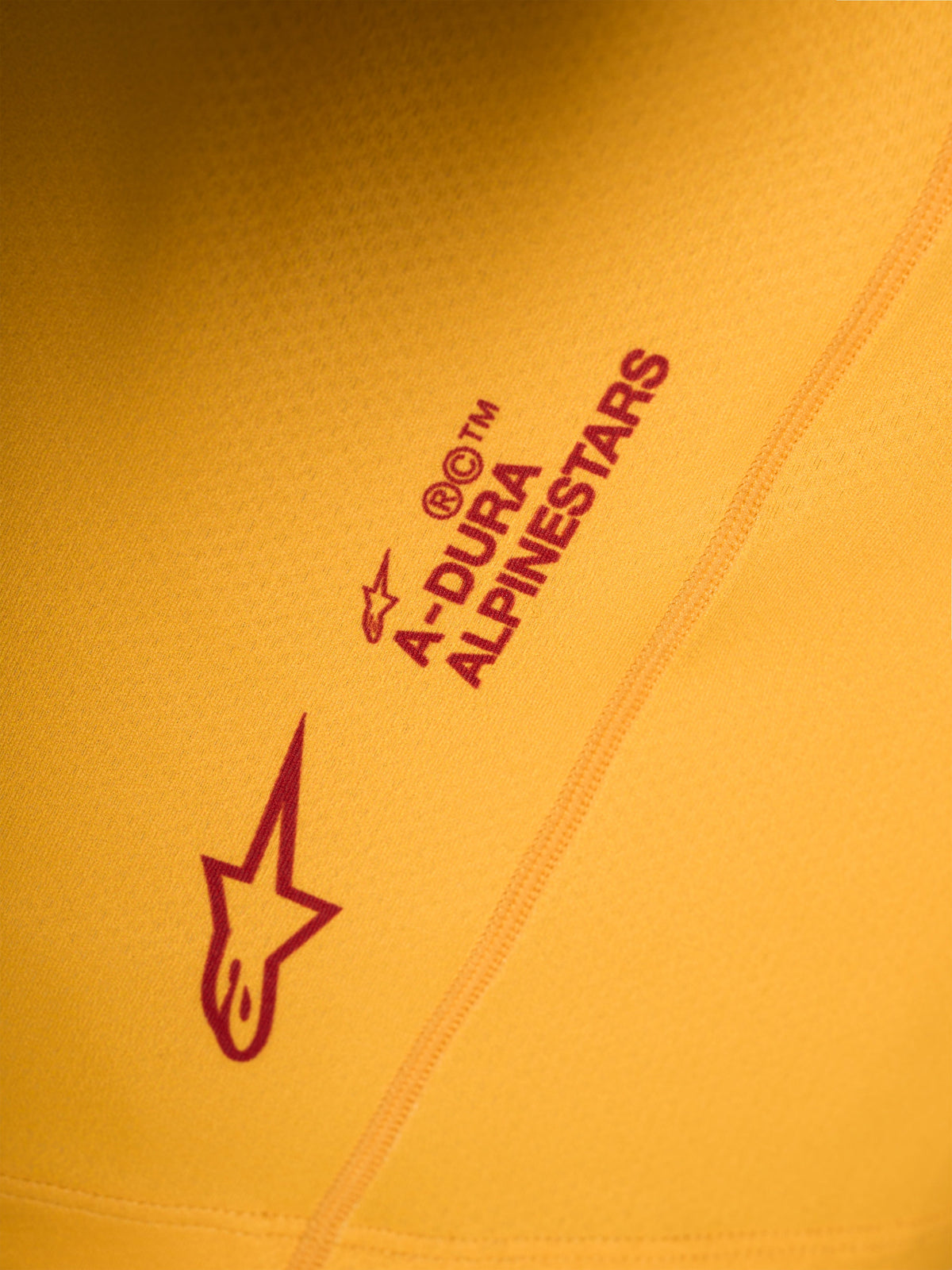 Pomarańczowa koszulka A-Dura Ride, detal z czerwonym logo Astars i napisem Alpinestars na elastycznej tkaninie.