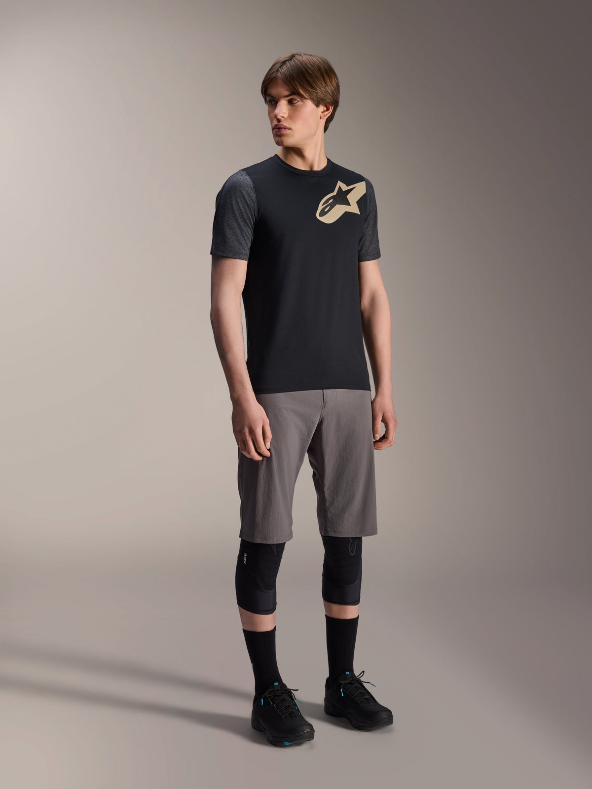 Męski model w koszulce Alpinestars A-Dura Lipan z krótkim rękawem w kolorze czarnym, z ciemnoszarymi rękawami i beżowym logo Astars na piersi, w zestawie z szarymi szortami rowerowymi, czarnymi ochraniaczami na kolana i czarnymi butami, kompletny strój do jazdy na rowerze górskim