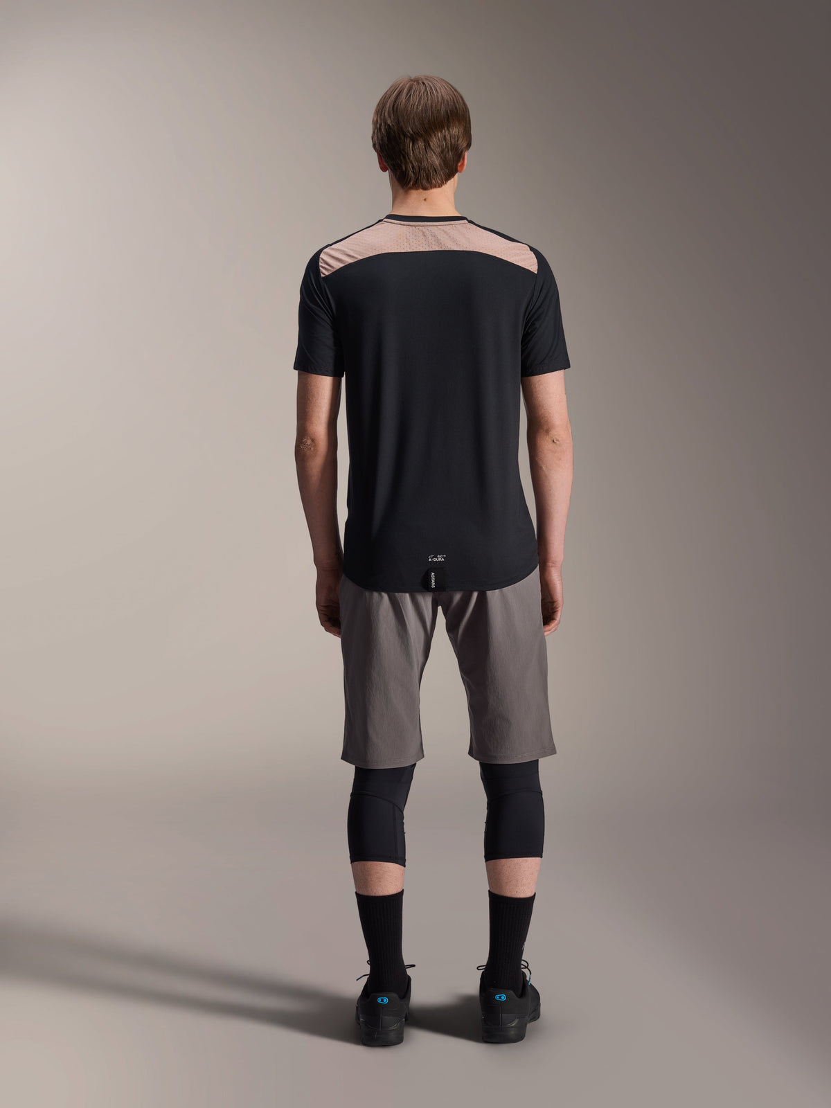 Alpinestars A-Dura Lipan Jersey - Short Sleeve, czarna z beżowym panelem na górze pleców, widok z tyłu pokazujący przedłużony tył, noszona z szarymi szortami MTB i czarnymi ochraniaczami kolan, wydajna odzież do jazdy na rowerze górskim