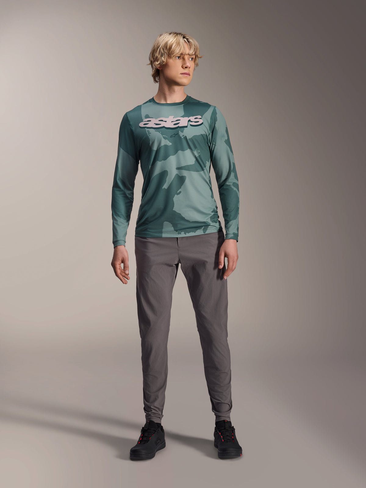 Alpinestars A-Dura Hollow Camo Long Sleeve Jersey, koszulka z długim rękawem, kolor Silver Pine Camo z zielonym wzorem moro, duże białe logo „astars” na klatce piersiowej, noszona z szarymi spodniami technicznymi i czarnymi butami rowerowymi, wysokiej jakości odzież MTB