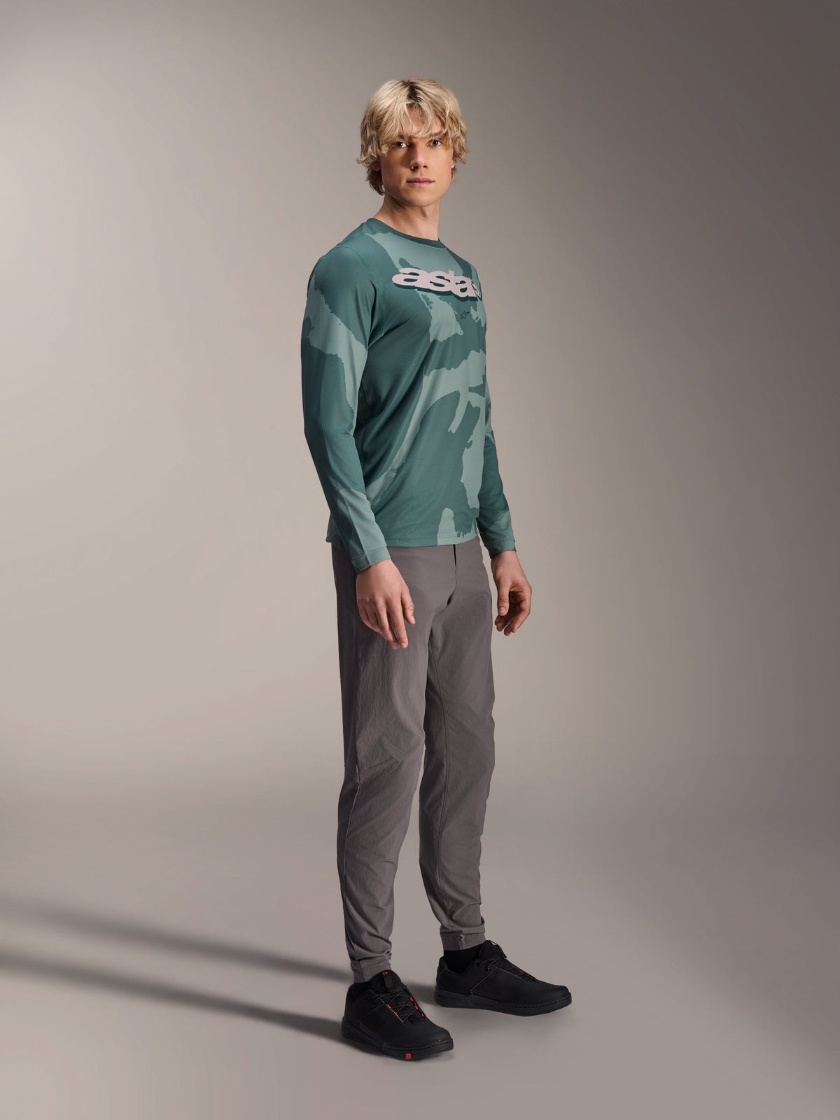 Alpinestars A-Dura Hollow Camo Long Sleeve Jersey, Silver Pine Camo, koszulka z długim rękawem noszona przez modela w szarych spodniach i czarnych butach, zielony wzór kamuflażu, wyraźne białe logo ASTARS na piersi, wysokiej klasy odzież MTB