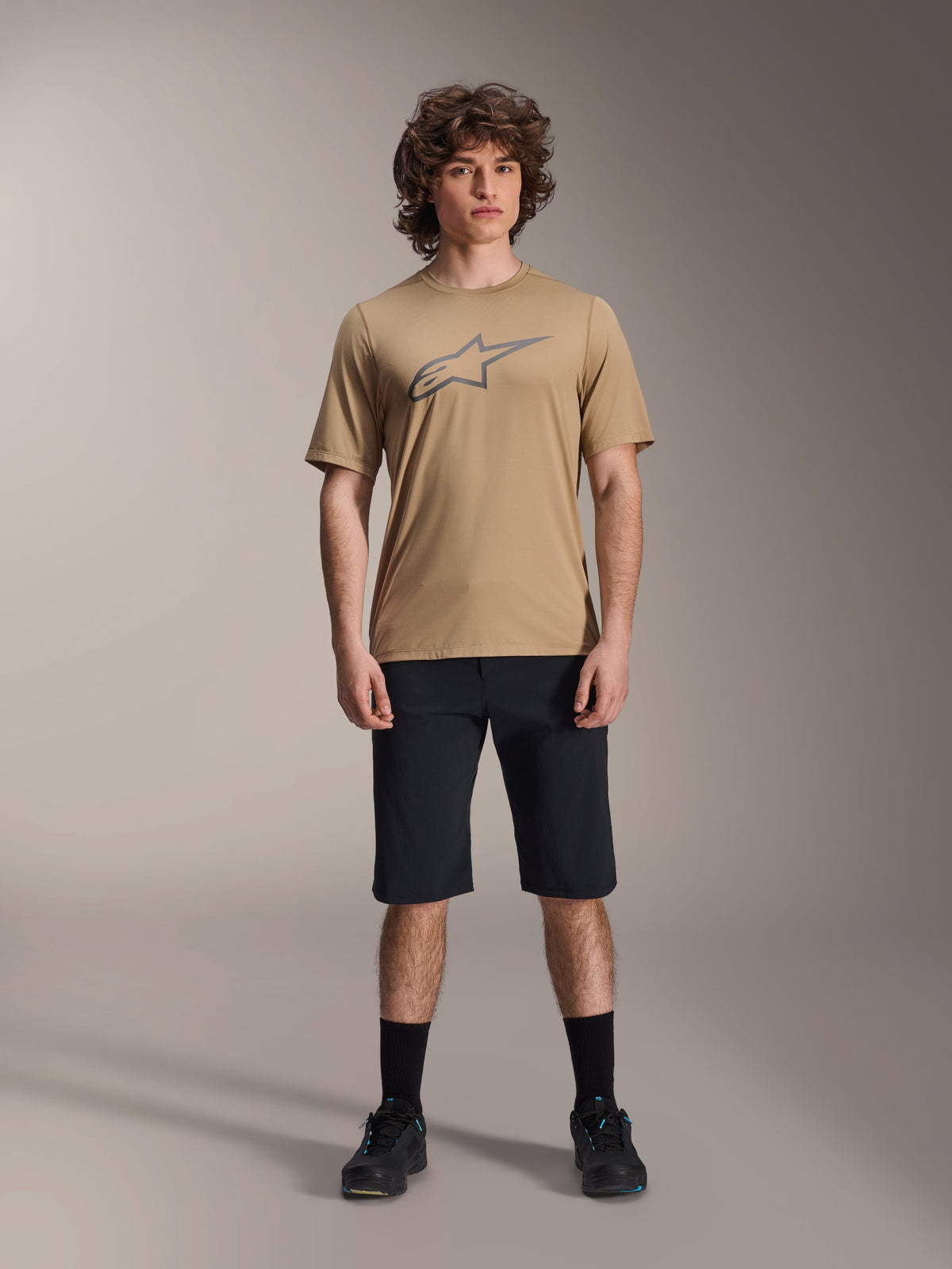 Alpinestars A-Dura Astar Short Sleeve Jersey, jasnobrązowa koszulka z krótkim rękawem, zestawiona z czarnymi szortami i czarnymi butami, duże konturowe logo Astar na piersi, techniczny materiał żakardowy do jazdy MTB