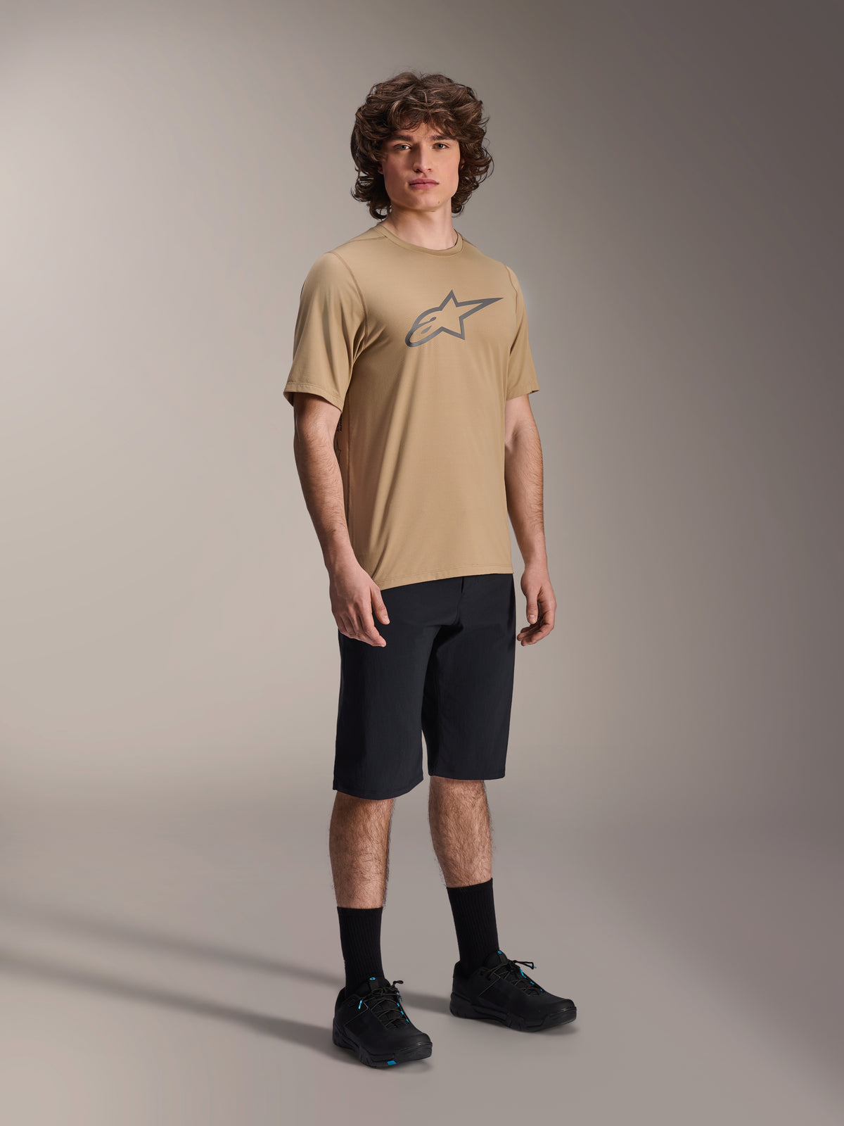 Spodenki Alpinestars A-Dura, czarne spodenki rowerowe MTB, prezentowane przez modela w beżowej koszulce z krótkim rękawem z logo Alpinestars, czarne skarpetki i czarne buty, wytrzymała odzież MTB do jazdy terenowej