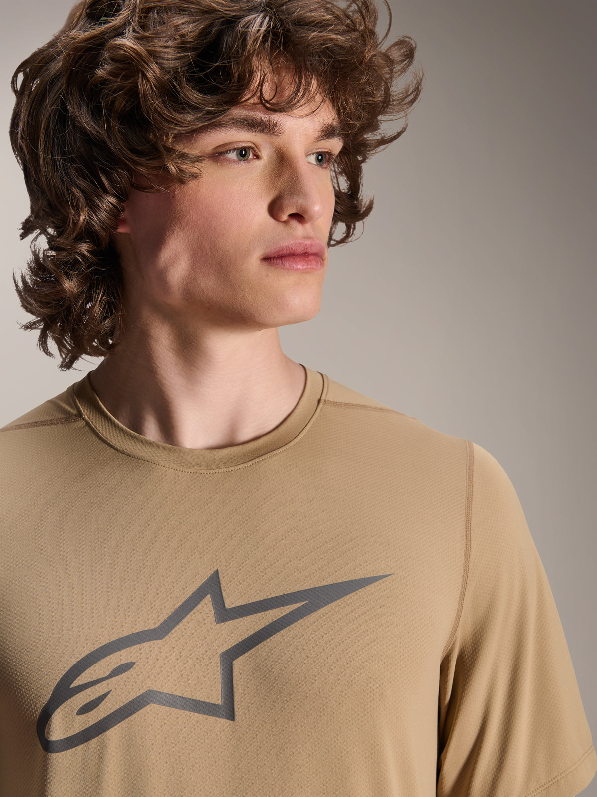 Model w koszulce Alpinestars A-Dura Astar z krótkim rękawem, jasnobrązowa koszulka MTB z szarym logo gwiazdy na piersi, odprowadzający wilgoć materiał żakardowy, casualowy styl rowerowy