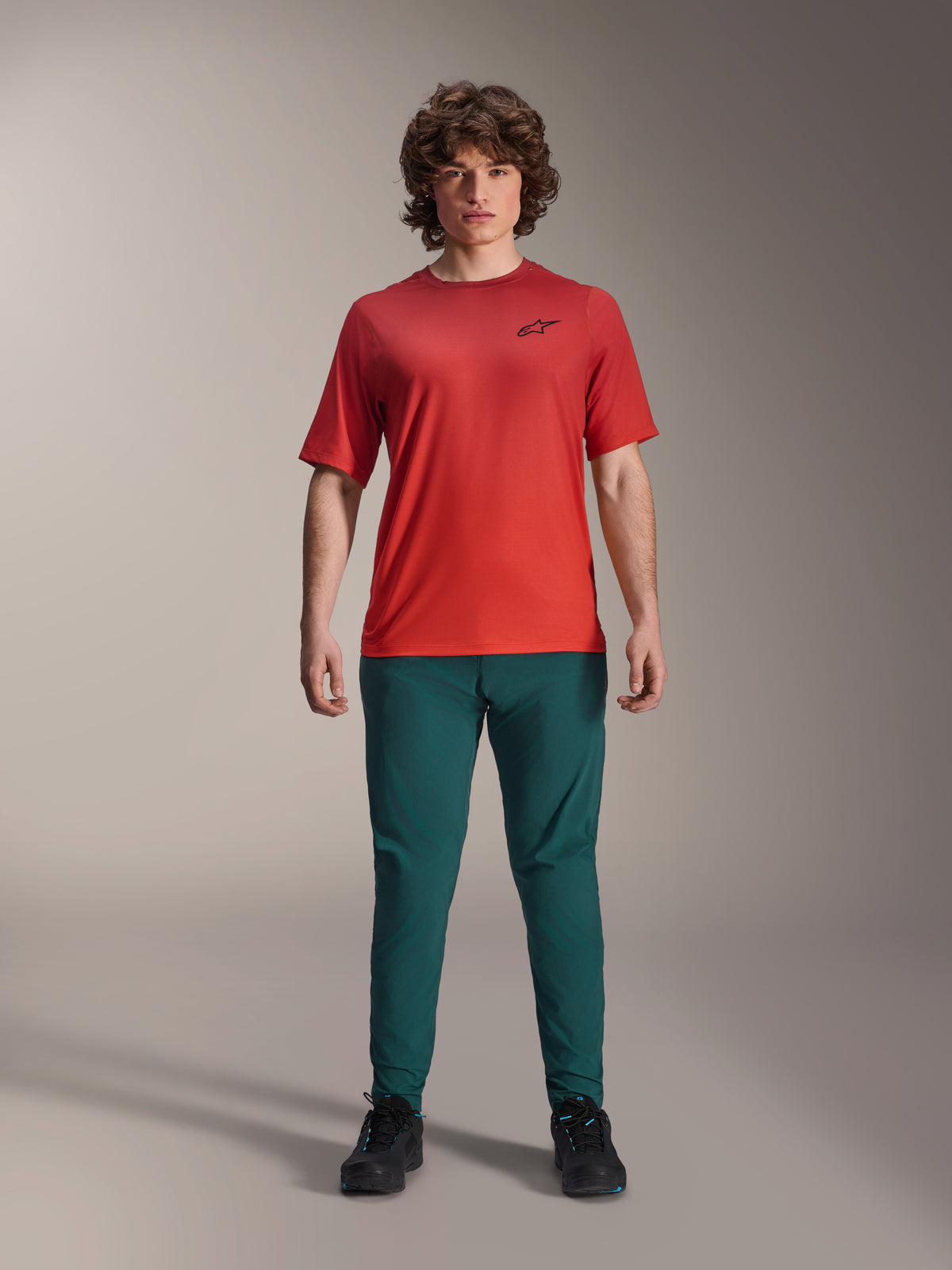 Spodnie Alpinestars A-Dura w kolorze Dark Pine green zestawione z czerwoną koszulką A-Dura Rise z krótkim rękawem, model pozujący w pełnym stroju MTB, swobodne dopasowanie typu performance, wytrzymała odzież rowerowa