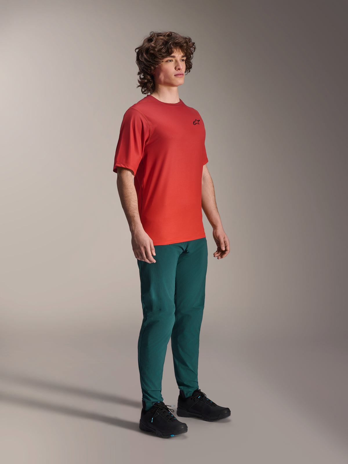 Model ubrany w spodnie Alpinestars A-Dura w kolorze Dark Pine, w połączeniu z czerwoną koszulką z krótkim rękawem A-Dura Rise i czarnymi butami MTB, sportowa odzież do kolarstwa górskiego w studio