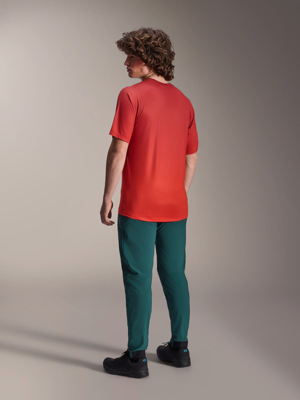 Model widziany od tyłu w spodniach Alpinestars A-Dura w kolorze ciemnozielonym Dark Pine oraz czerwonej koszulce z krótkim rękawem A-Dura Rise, regularny krój, elastyczne mankiety, profesjonalna odzież MTB do jazdy w terenie
