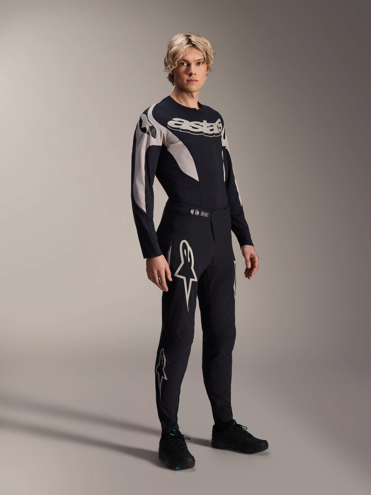 Męski model w stroju Alpinestars A-Supra Melt, koszulka z długim rękawem i spodnie, czarny zestaw z jasnoszarymi zakrzywionymi wzorami, białe logo Astars na nogawkach i napis na klatce piersiowej, profesjonalny strój do wyścigów MTB