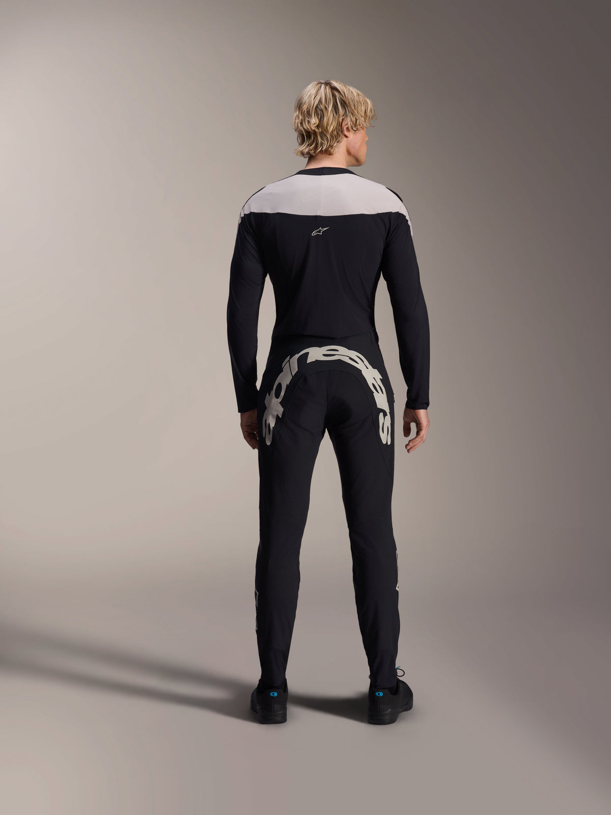 Alpinestars A-Supra Melt Pants, czarne, widok z tyłu pokazujący duży biały napis Alpinestars na tylnym karczku, zestawione z pasującą koszulką z długim rękawem i czarnymi butami rowerowymi, wysokiej jakości odzież MTB do jazdy w terenie