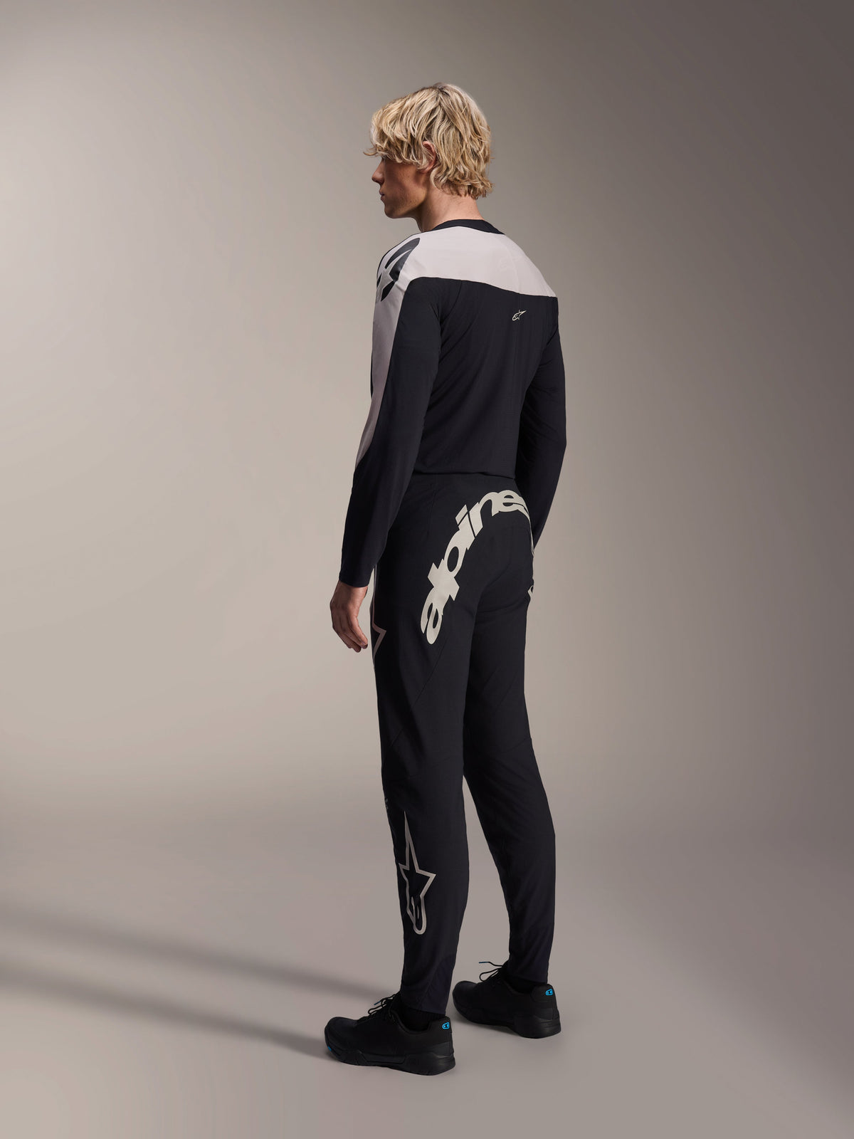 Alpinestars A-Supra Melt Pants, spodnie MTB, czarne, z dużym białym napisem Alpinestars na tylnym karczku i logo gwiazdy na nogawce, noszone przez modela z dopasowaną koszulką z długim rękawem i czarnymi butami, widok z tyłu z boku