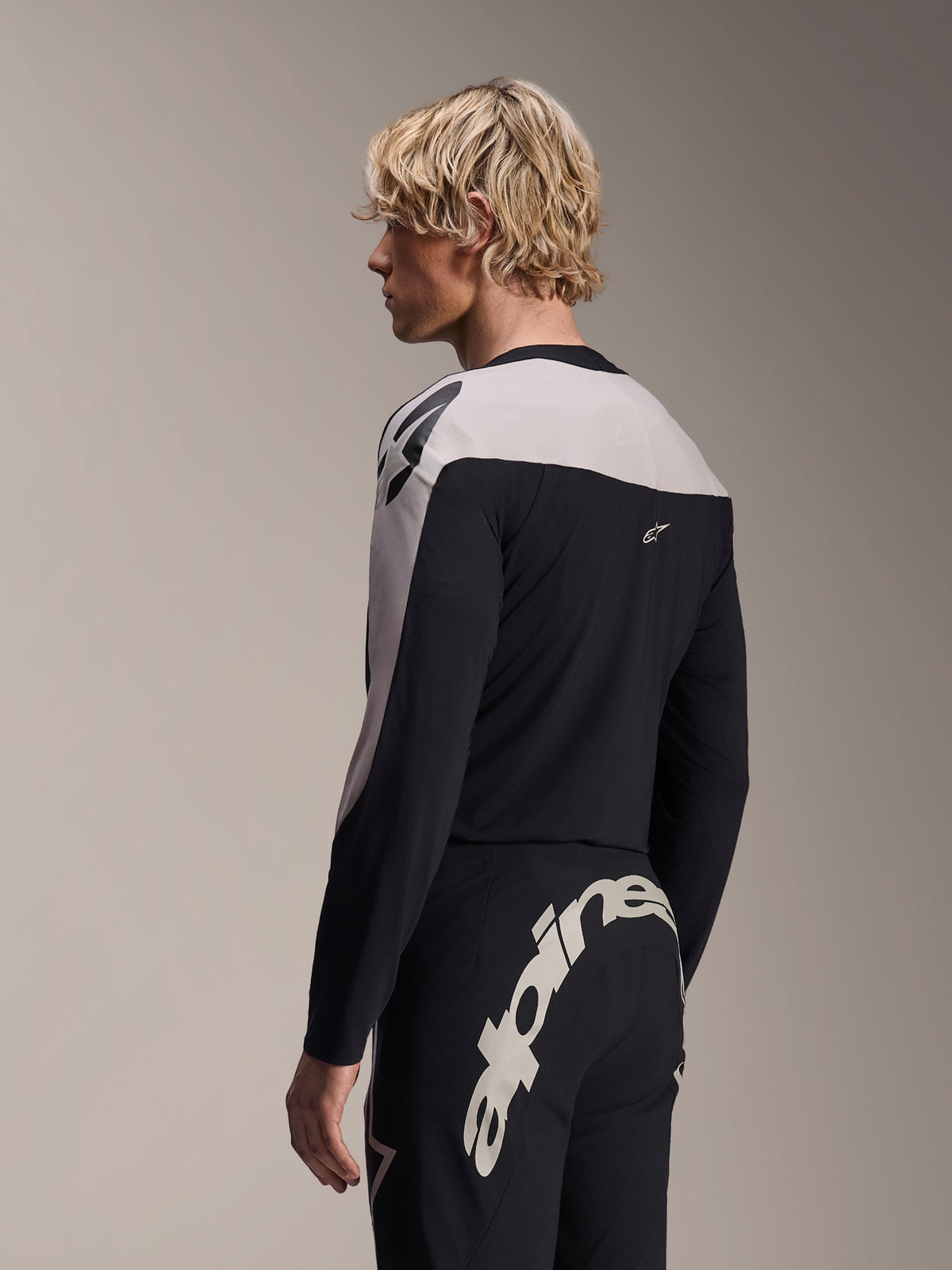 Model w koszulce Alpinestars A-Supra Melt z długim rękawem i spodniach A-Supra Melt, czarno-jasnoszary strój wyścigowy MTB, z dużym białym napisem Alpinestars na karczku z tyłu spodni