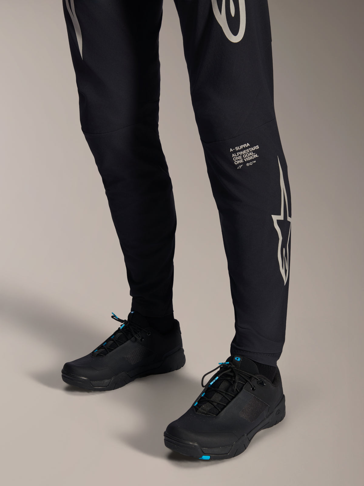 Alpinestars A-Supra Melt Pants, czarne spodnie rowerowe MTB, z białym napisem i logo Alpinestars na nogawce, zestawione z czarnymi butami rowerowymi z niebieskimi akcentami, wyczynowa odzież terenowa