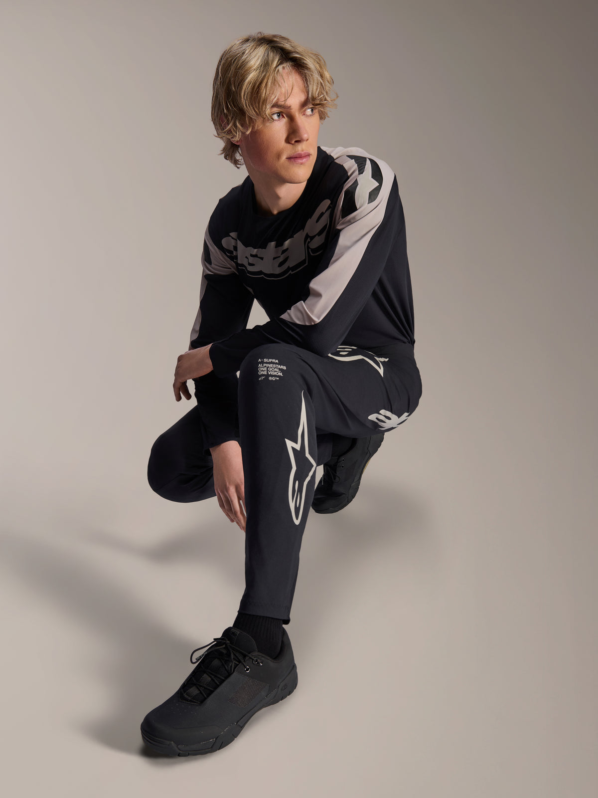 Alpinestars A-Supra Melt Pants i koszulka z długim rękawem, czarno-szare, z wyraźnym białym brandingiem i logo gwiazdy, model w pozycji kucającej, dopasowany zestaw wyścigowy MTB