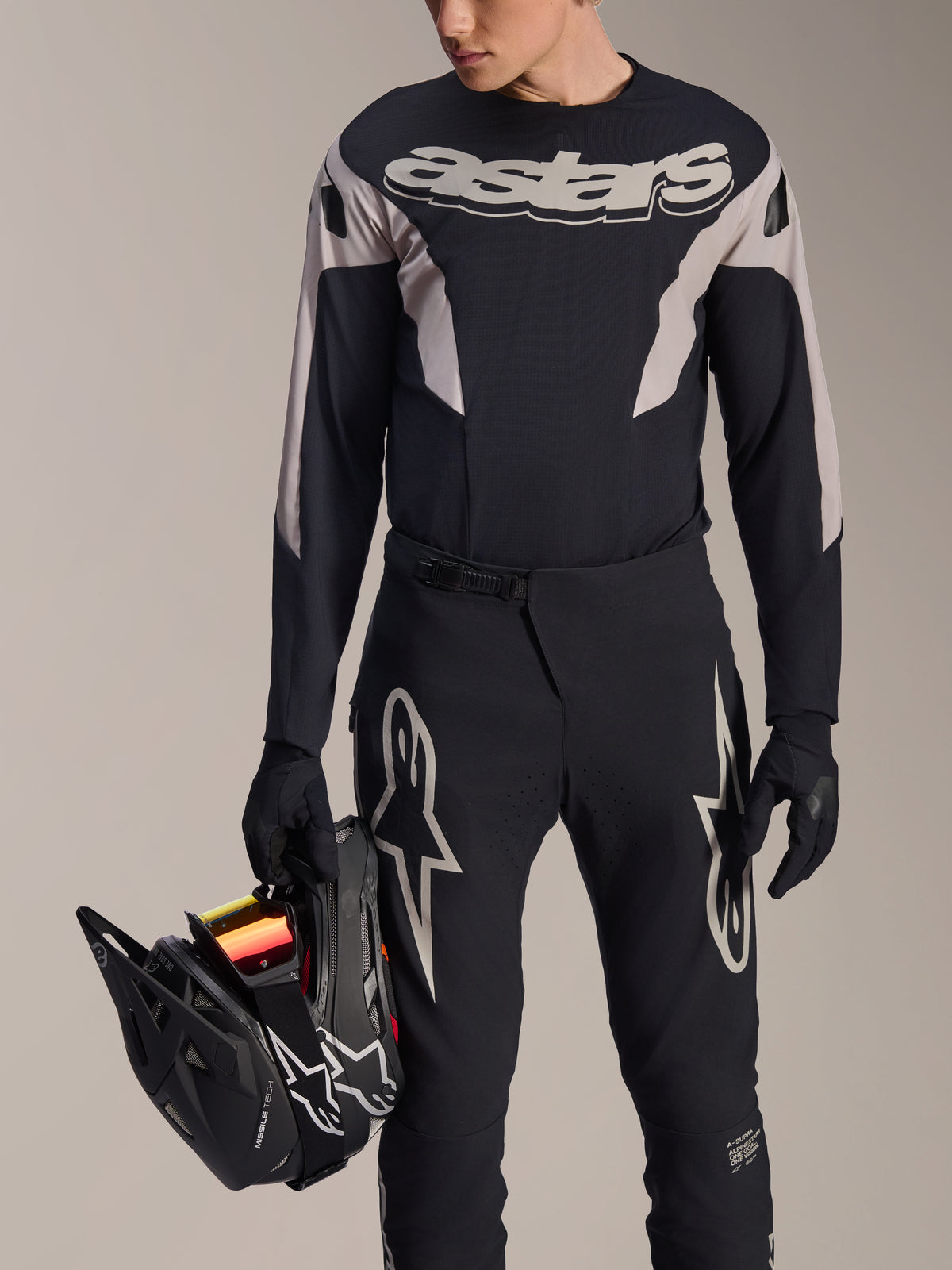 Zestaw wyścigowy MTB Alpinestars A-Supra Melt, koszulka z długim rękawem i spodnie, czarno-szary z białym logo Astars, model trzymający czarny kask full-face i mający na sobie pasujące rękawiczki, wyczynowa odzież do downhillu i enduro