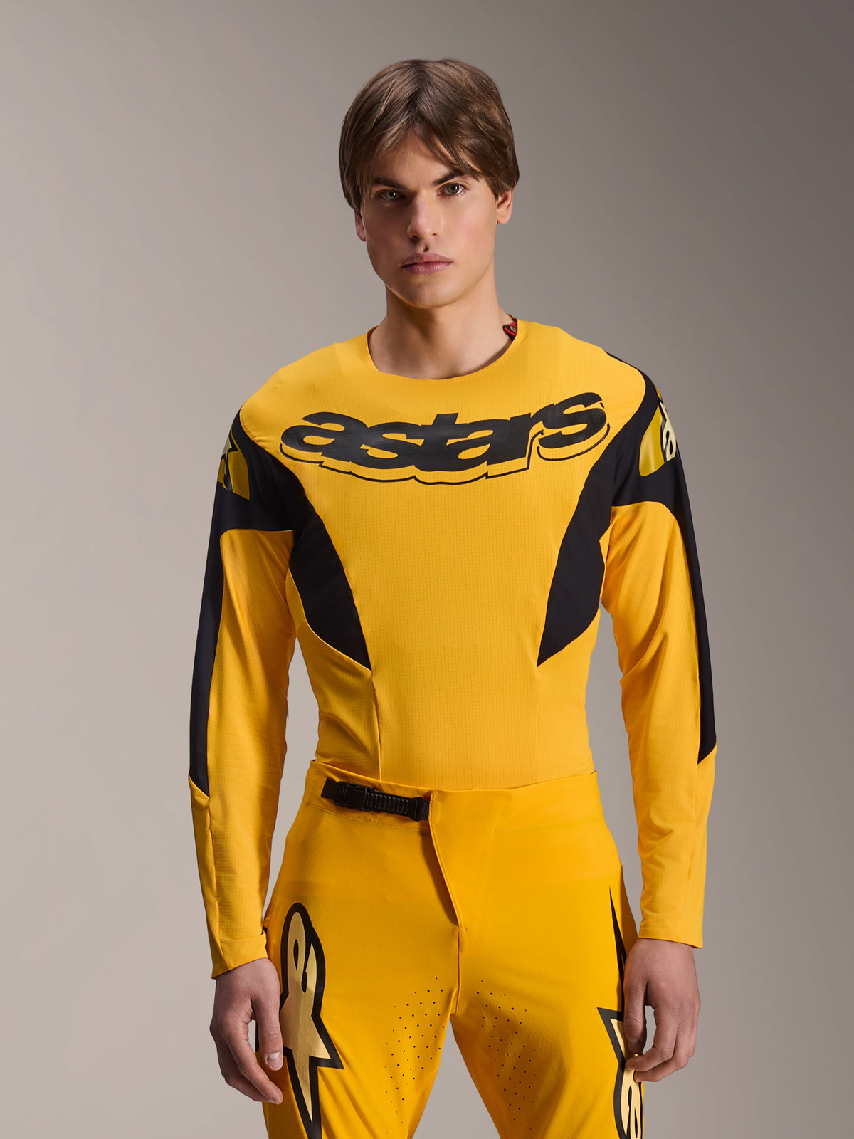 Alpinestars Limited Edition A-Supra Melt Gold Jersey, koszulka MTB z długim rękawem, ciemnożółta z czarnymi akcentami, prezentowana przez modela z dopasowanymi spodniami, z dużym czarnym logo „astars” na piersi i sportowym, wyczynowym designem
