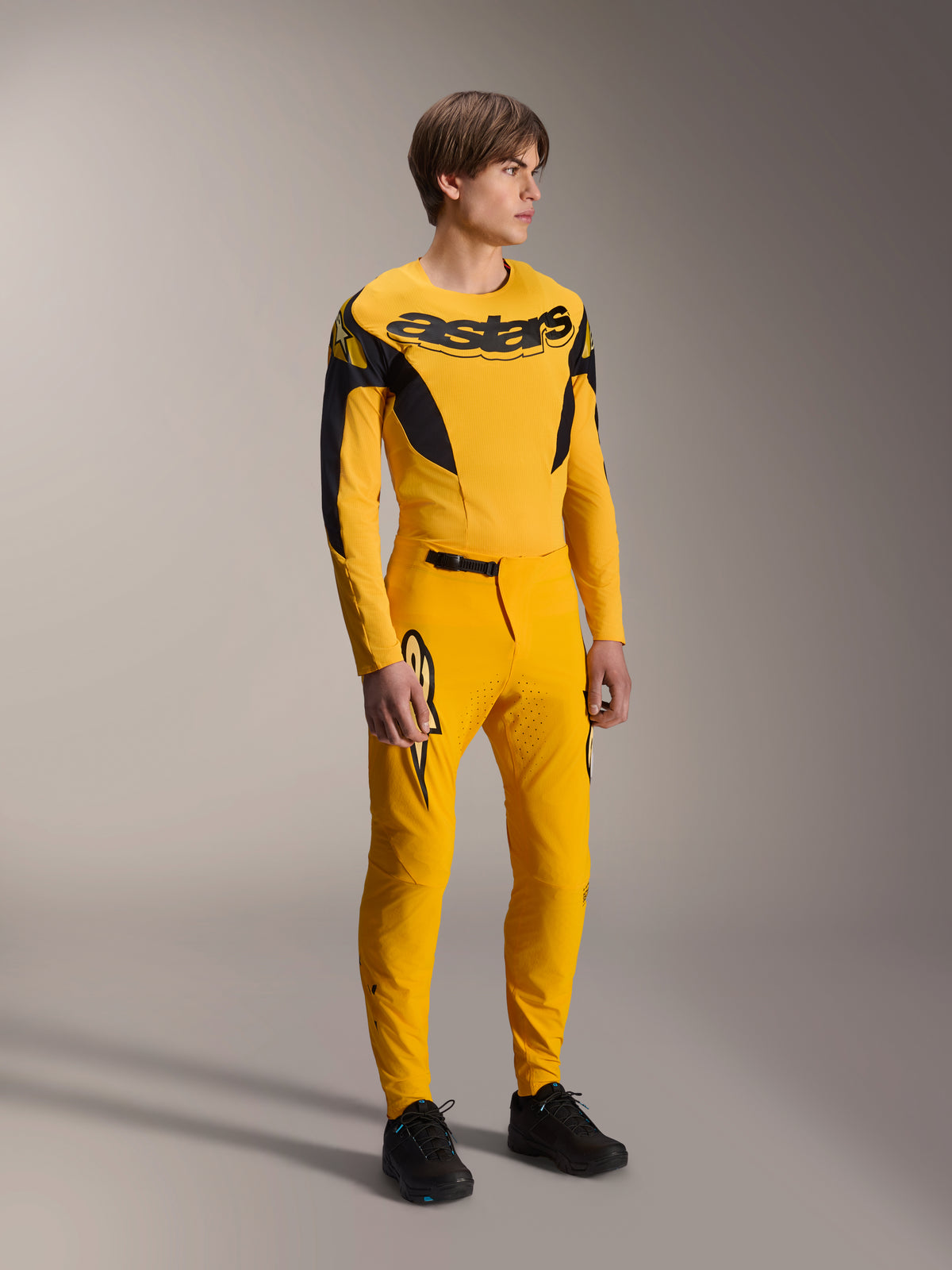 Męski model w zestawie Alpinestars Limited Edition A-Supra Melt Gold, żółta koszulka MTB z długim rękawem i wyraźnym czarnym logo na piersi, pasujące żółte spodnie z laserowo wycinaną wentylacją i czarnymi akcentami, kompletny zestaw wyścigowy do jazdy na rowerze górskim.