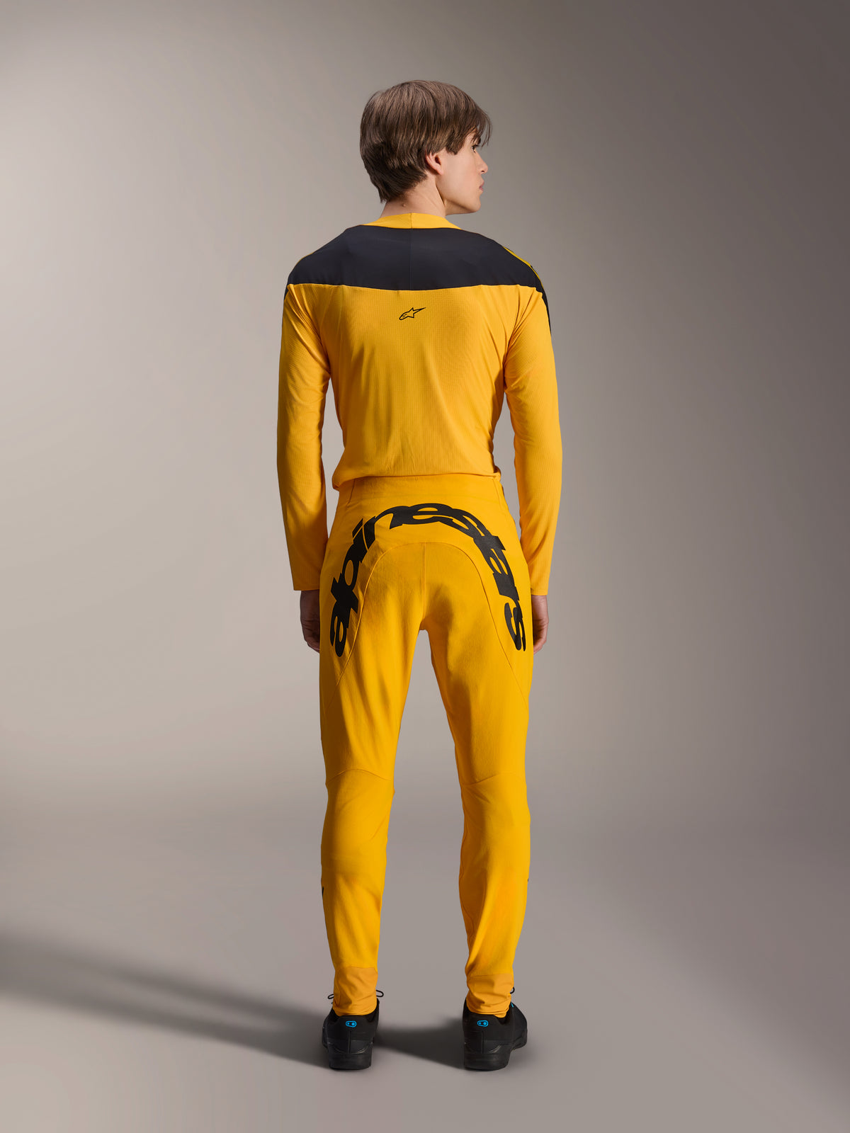 Widok z tyłu modelu ubranego w zestaw Alpinestars Limited Edition A-Supra Melt Gold Gear-Set, składający się z ciemnożółtej koszulki z długim rękawem z czarnym karczkiem na ramionach oraz dopasowanych ciemnożółtych spodni z dużym czarnym napisem Alpinestars na tylnym karczku, w połączeniu z czarnymi butami.