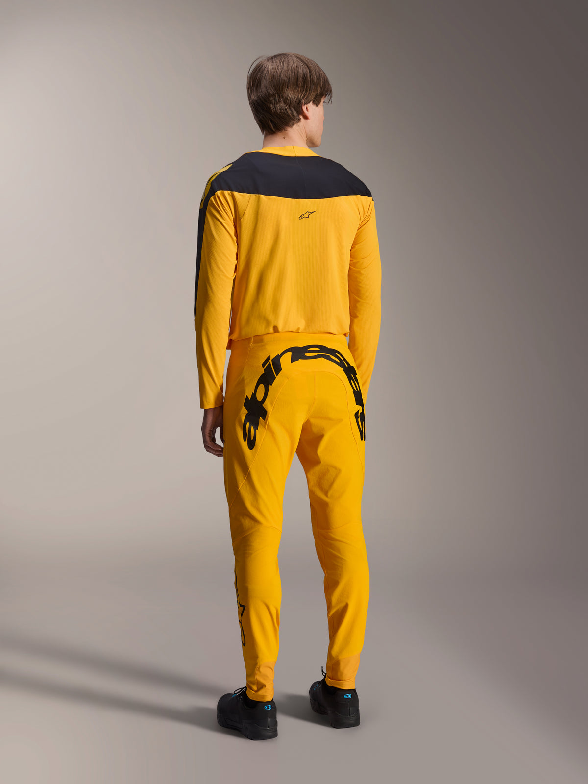 Alpinestars Limited Edition A-Supra Melt Gold Gear-Set, koszulka i spodnie MTB, kolor ciemnożółty i czarny, widok z tyłu z dużym napisem Alpinestars na siedzeniu i czarnym karczkiem na koszulce, wyczynowa odzież do kolarstwa górskiego