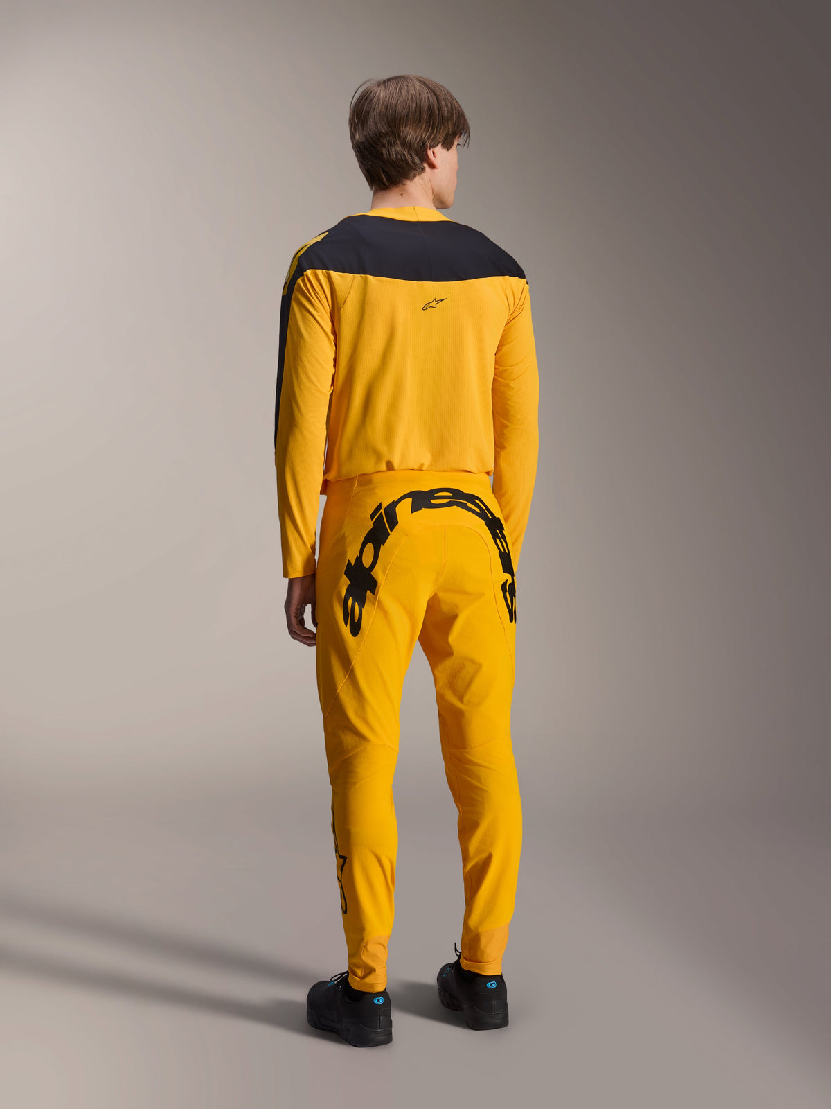 Koszulka Alpinestars Limited Edition A-Supra Melt Gold, koszulka MTB z długim rękawem, ciemnożółta z czarnym panelem na ramionach, w zestawie z dopasowanymi żółtymi spodniami z wyraźnym czarnym brandingiem z tyłu, widok z tyłu wyczynowego stroju kolarskiego
