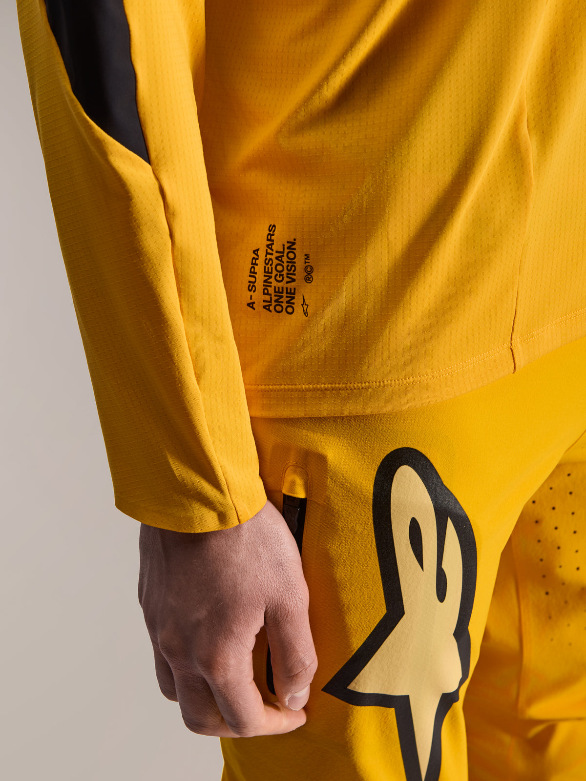 Alpinestars Limited Edition A-Supra Melt Gold Gear-Set, zbliżenie na ciemnożółtą koszulkę z długim rękawem i pasujące spodnie, z napisem „One Goal. One Vision.” oraz dużym logo Astars, mikroperforowana tkanina techniczna do wyścigów MTB