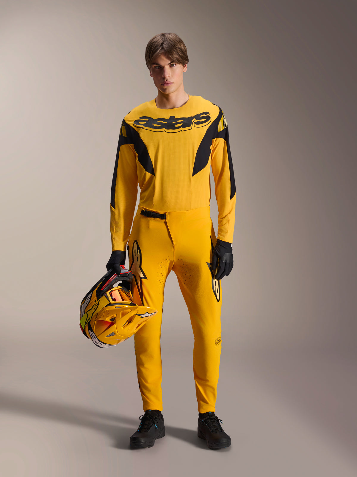 Model w zestawie MTB Alpinestars Limited Edition A-Supra Melt Gold, żółta koszulka z długim rękawem z czarnym logo „astars”, żółte spodnie z czarnymi akcentami, żółto-czarny kask, czarne rękawice i buty, profesjonalna odzież do kolarstwa górskiego