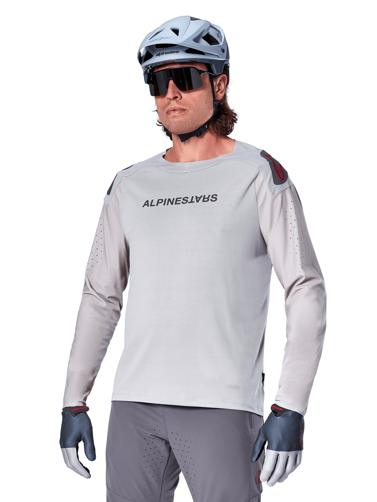 Alpinestars A-Aria Polartec Switch Jersey Long Sleeve, jasnoszara koszulka rowerowa z długim rękawem, czarne logo na klatce piersiowej i czerwony akcent na ramieniu, laserowo perforowane rękawy dla lepszej wentylacji, noszona z jasnoniebieskim kaskiem, okularami przeciwsłonecznymi i szarymi spodniami, profesjonalna odzież MTB na szlaki