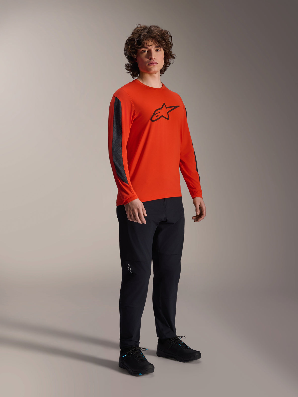Mężczyzna w koszulce Alpinestars A-Dura Insert Astar z długim rękawem, kolor Spicy Orange z czarnym logo Astar na klatce piersiowej, ciemnoszare wstawki na rękawach, czarne spodnie, profesjonalna odzież rowerowa MTB