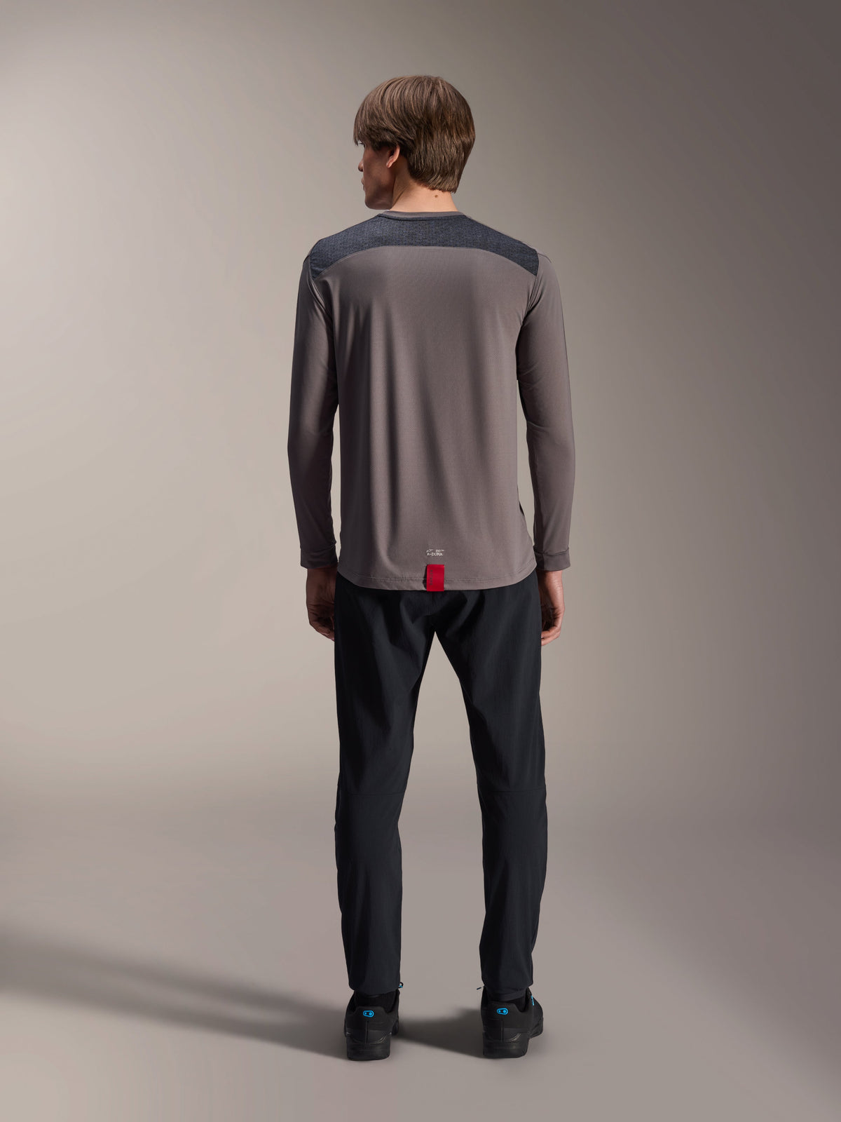 Alpinestars A-Dura Insert Astar Long Sleeve Jersey in Dark Gray, rear view, featuring charcoal shoulder panels and a red tag at the hem, worn with black pants and shoes, mountain bike performance apparel