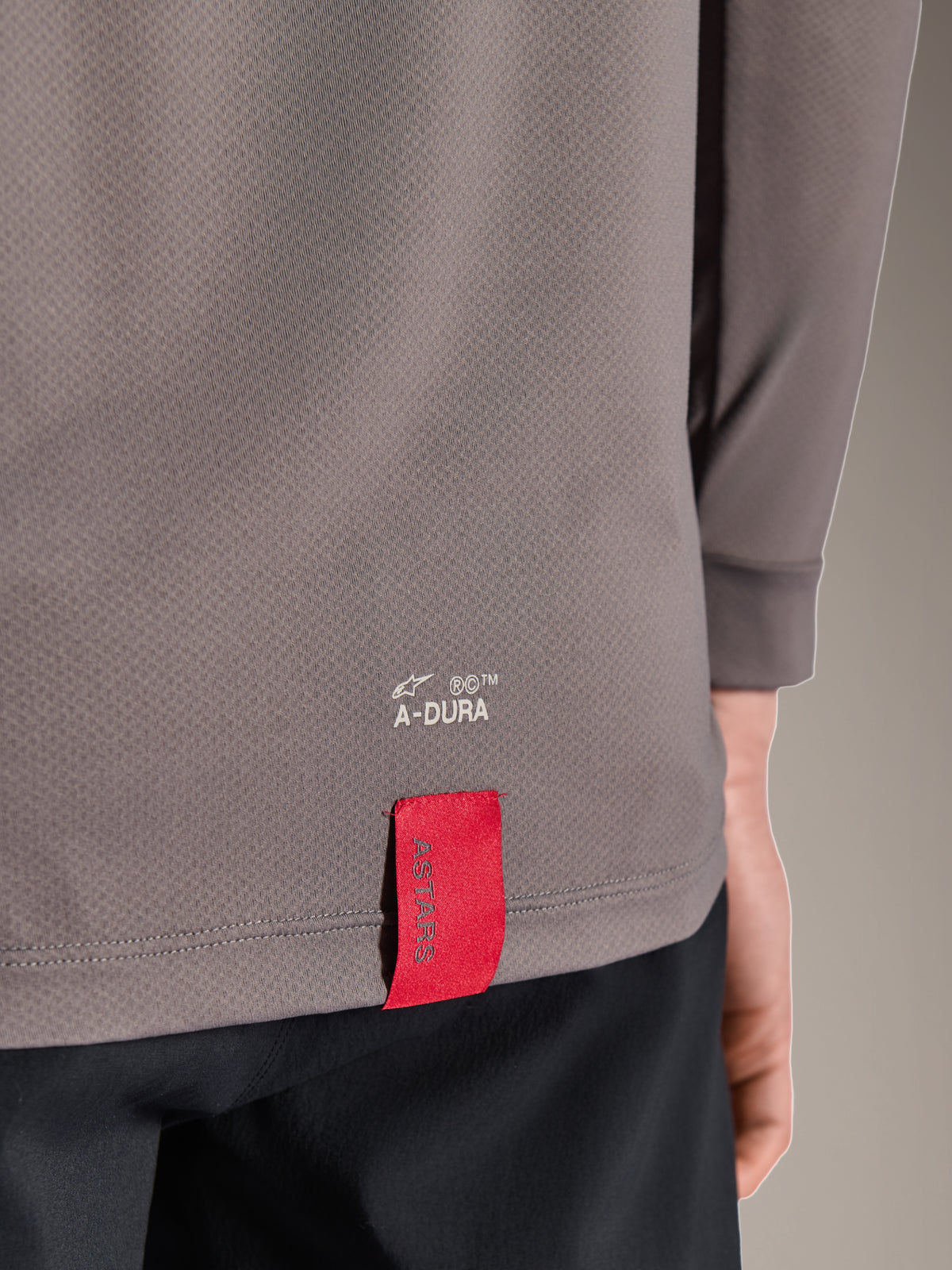 Alpinestars A-Dura Insert Astar Jersey, Long Sleeve MTB Jersey, Dark Gray, close-up detail of lower back hem featuring white A-DURA branding and a signature red ASTARS fabric tag, textured performance fabric