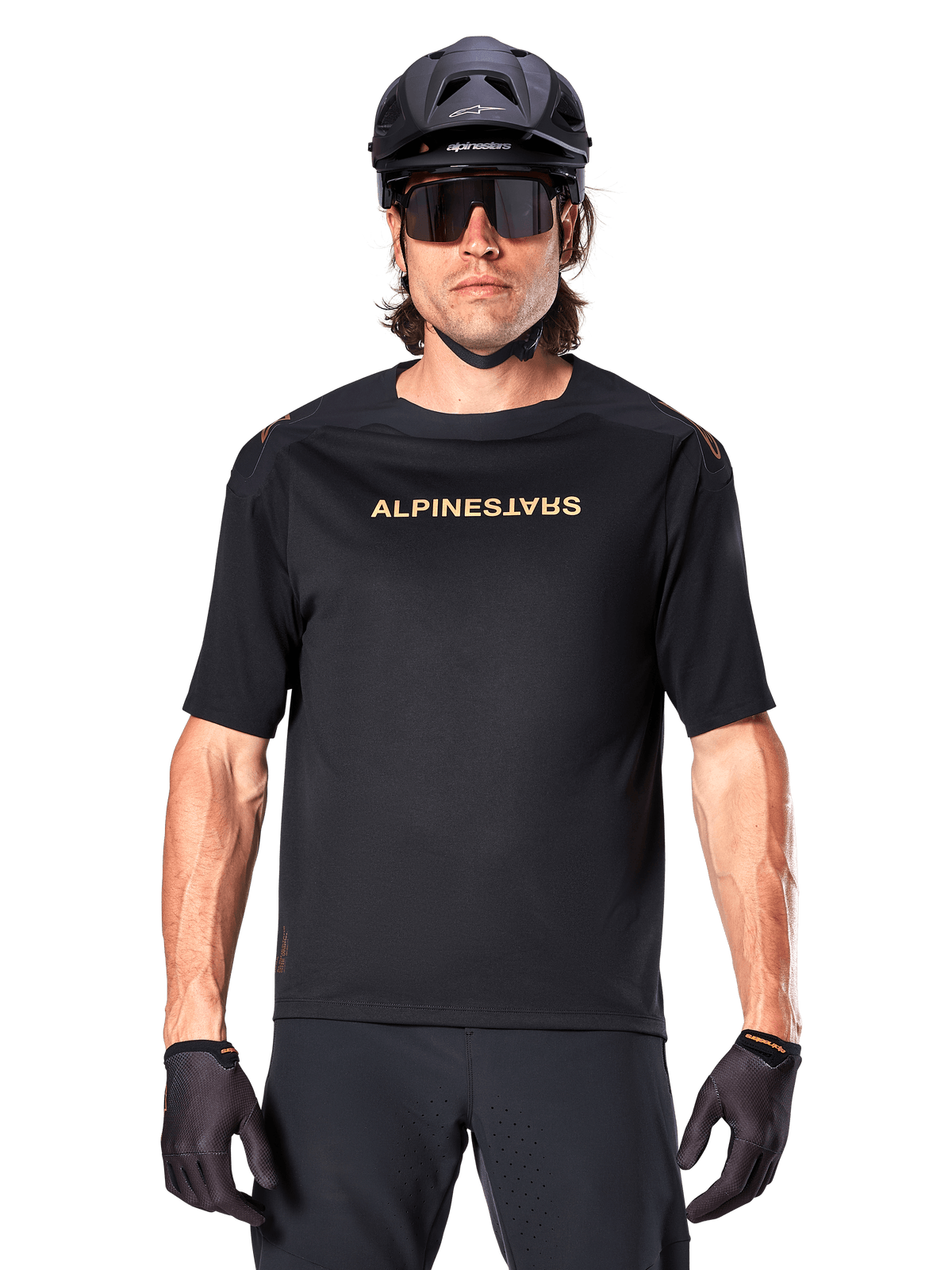 Rowerzysta górski w koszulce Alpinestars A-Aria Polartec Switch z krótkim rękawem w kolorze czarnym, ze złotym brandingiem na klatce piersiowej, w zestawie z czarnym kaskiem MTB, ciemnymi okularami sportowymi i pasującymi rękawiczkami, odzież do jazdy terenowej.