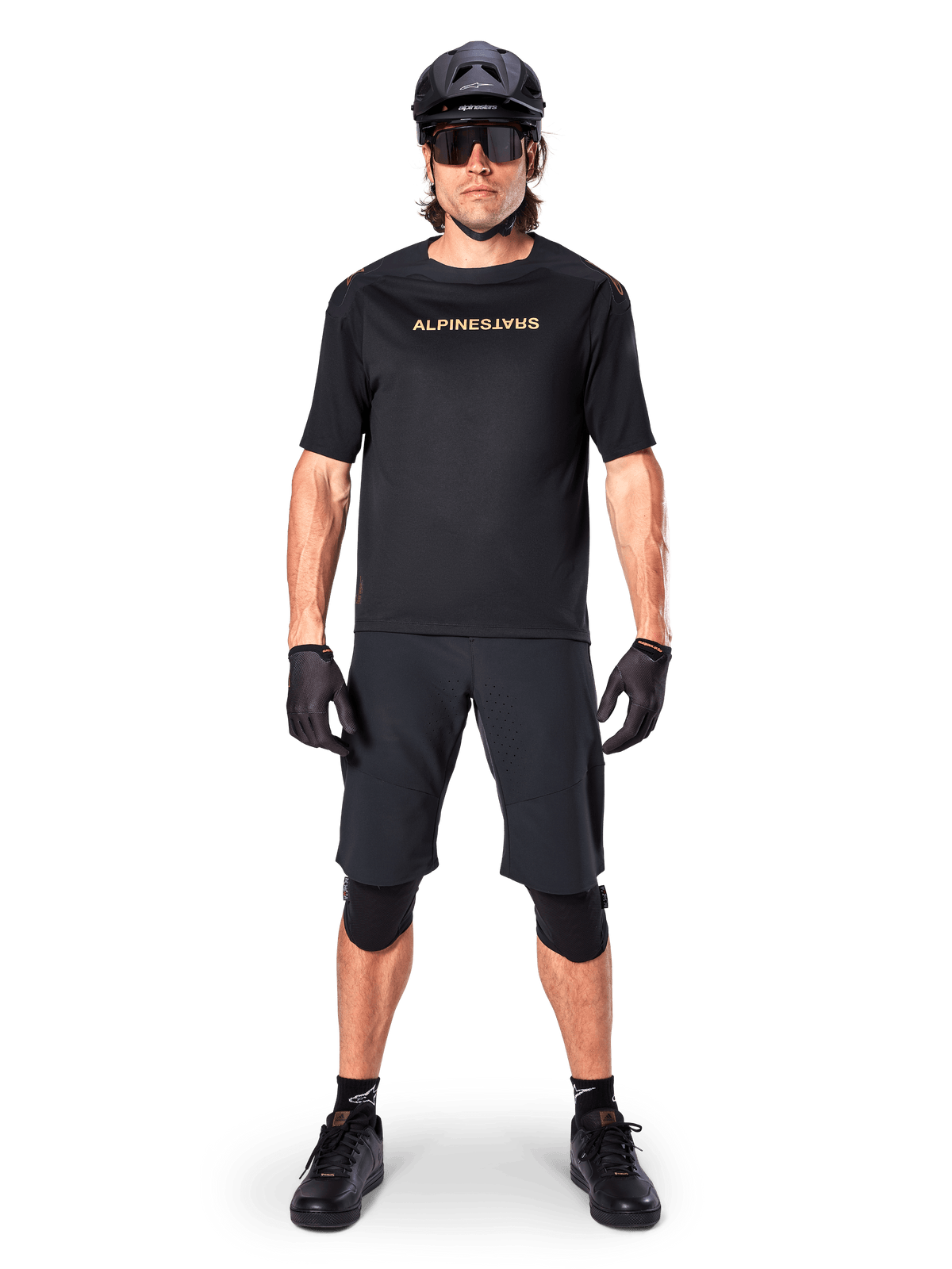 Alpinestars A-Aria Polartec Switch Jersey, koszulka MTB z krótkim rękawem, czarna z beżowymi napisami, noszona z dopasowanym czarnym ekwipunkiem do kolarstwa górskiego, w tym kaskiem, szortami i ochraniaczami na kolana, techniczna odzież rowerowa do jazdy w terenie