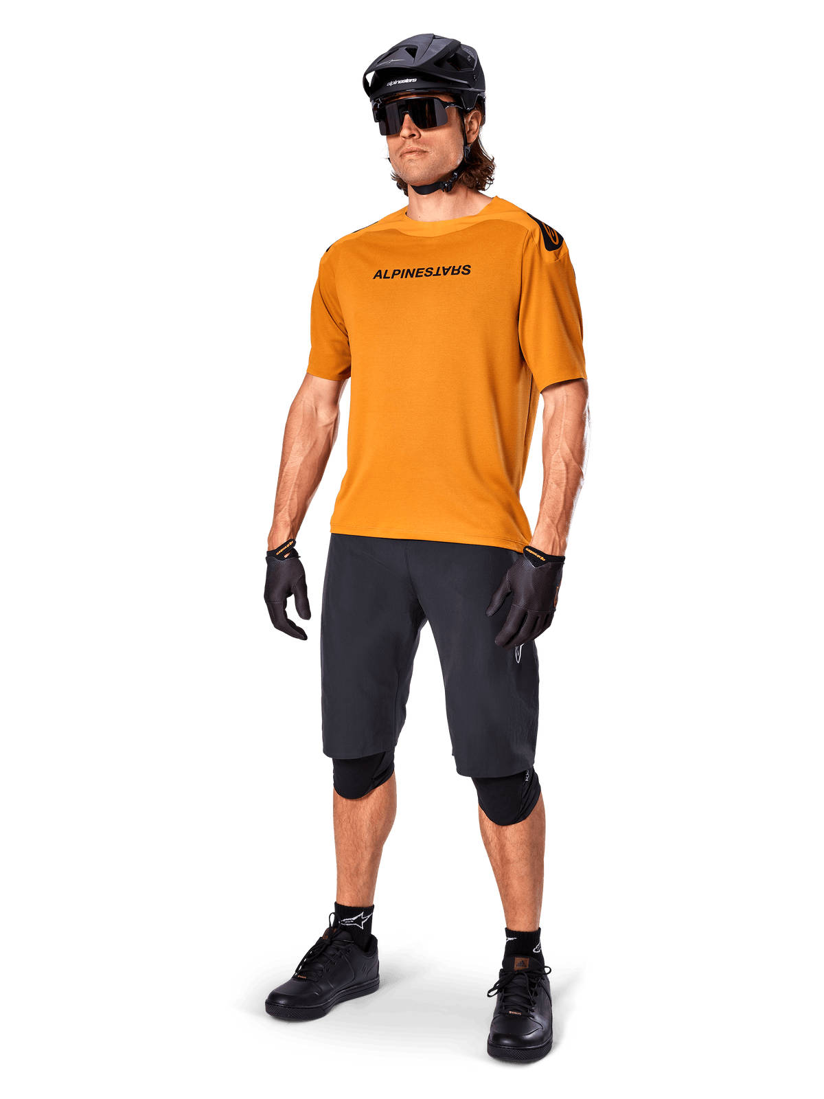 Rowerzysta w koszulce Alpinestars A-Aria Polartec Switch w kolorze Dark Gold, koszulka MTB z krótkim rękawem, zestawiona z czarnymi spodenkami rowerowymi, nakolannikami, czarnym kaskiem i rękawiczkami, profesjonalny strój do jazdy na rowerze górskim po szlakach