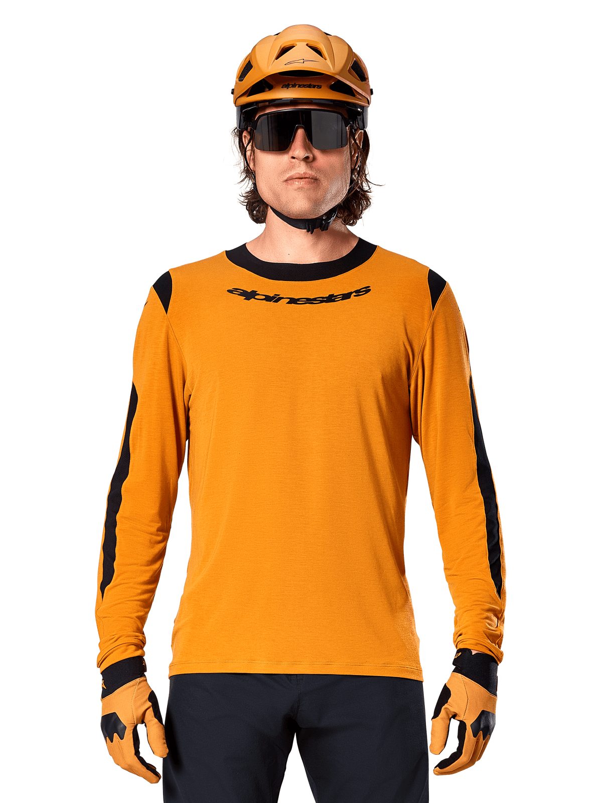 Męski model w koszulce z długim rękawem Alpinestars A-Dura Dri Wool w kolorze Dark Gold, dopasowany złoty kask MTB i rękawice, czarne panele z siatki na ramionach i rękawach, czarny napis Alpinestars na piersi, ciemne okulary przeciwsłoneczne i spodnie, techniczna odzież do kolarstwa górskiego
