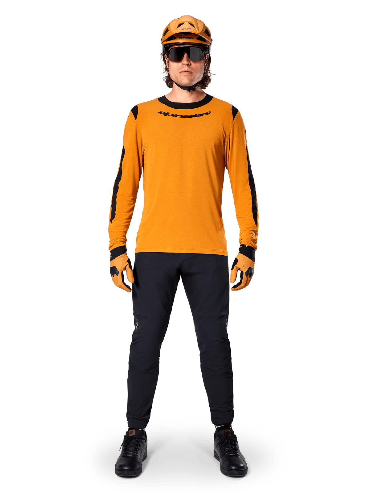 Rowerzysta górski w koszulce Alpinestars A-Dura Dri Wool Long Sleeve Jersey, ciemnozłota z czarnymi akcentami na kołnierzu i rękawach, czarne spodnie, pasujący złoty kask, rękawice i czarne okulary przeciwsłoneczne, profesjonalny sprzęt do kolarstwa górskiego
