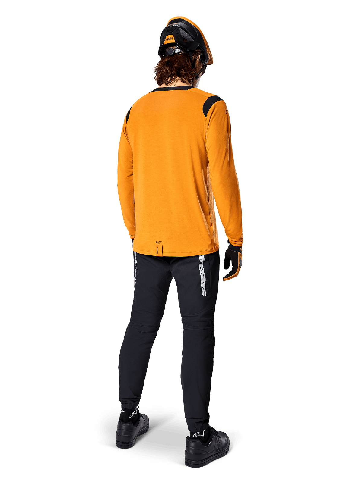 Rowerzysta górski widziany od tyłu w koszulce Alpinestars A-Dura Dri Wool z długim rękawem w kolorze Dark Gold, czarne spodnie techniczne z białym logo i dopasowany czarno-pomarańczowy kask, profesjonalna odzież do kolarstwa górskiego