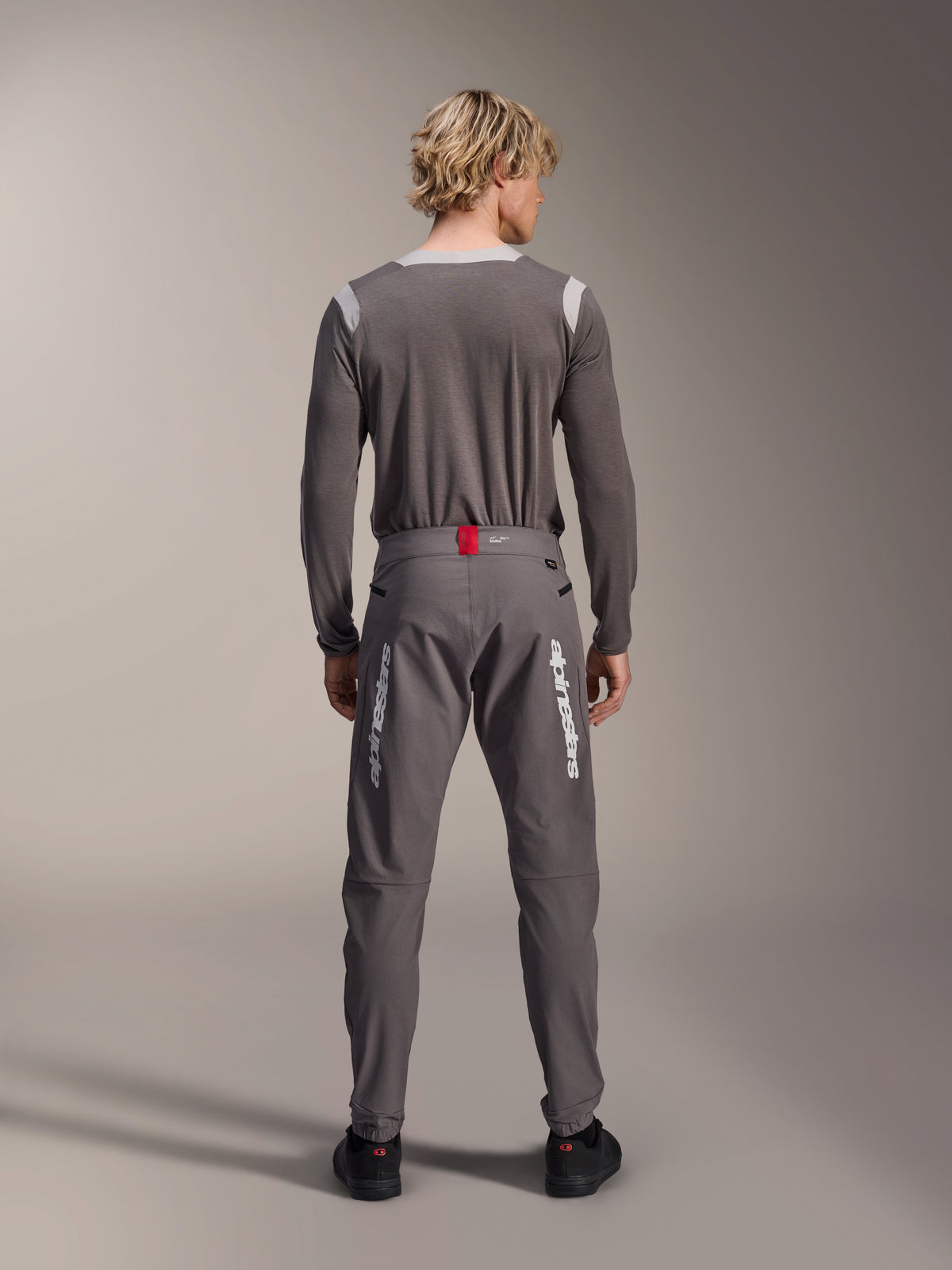 Model from behind wearing Alpinestars A-Dura Elite Pants in Dark Gray and a matching gray long-sleeve jersey, featuring white Alpinestars logos on the legs, a red accent on the waistband, and black shoes, professional MTB riding gear.