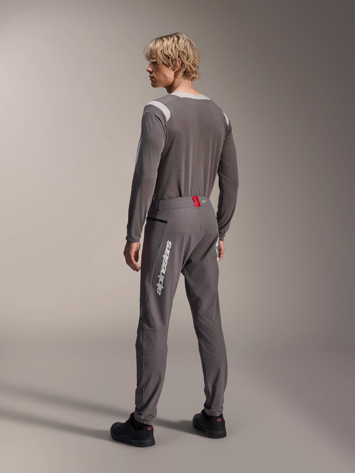 Model from a rear view wearing Alpinestars MTB gear, gray A-Dura Dri Wool Long Sleeve Jersey, Dark Gray A-Dura Elite Pants with white vertical branding and red waistband detail, coordinated cycling outfit for mountain biking