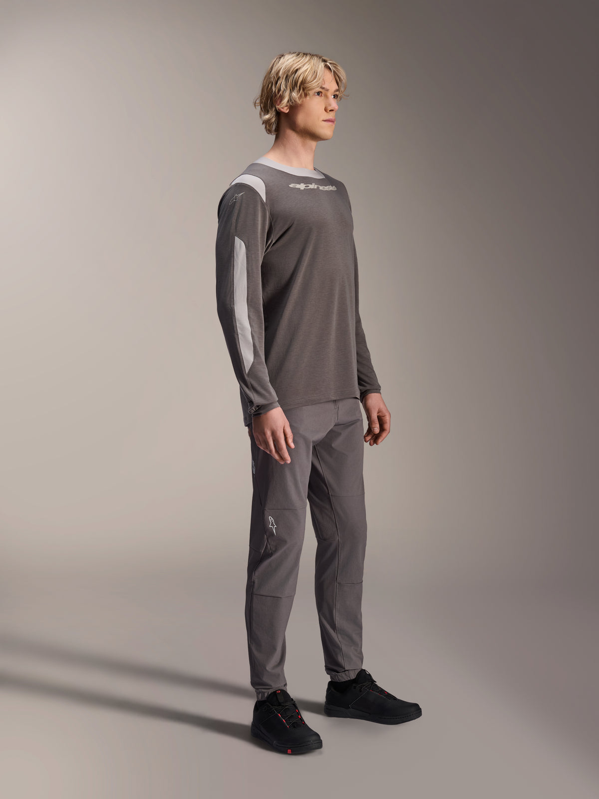 Męski model w koszulce Alpinestars A-Dura Dri Wool z długim rękawem w kolorze ciemnoszarym, zestawionej z pasującymi szarymi spodniami i czarnymi butami, jasnoszare panele z siateczki na ramionach i rękawach, techniczna odzież MTB do jazdy terenowej