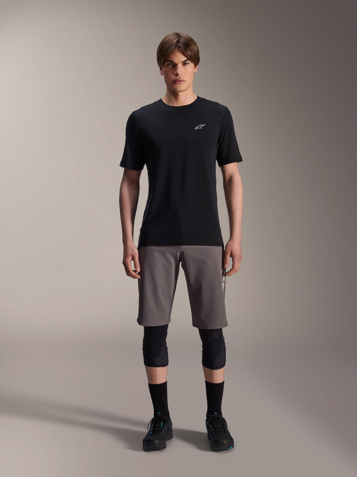 Męski model ubrany w spodenki Alpinestars A-Dura Elite w kolorze ciemnoszarym i koszulkę z krótkim rękawem A-Dura Dri-Wool w kolorze czarnym, w połączeniu z czarnymi ochraniaczami kolan i butami rowerowymi, techniczna odzież do rowerów górskich zapewniająca wydajność w terenie.