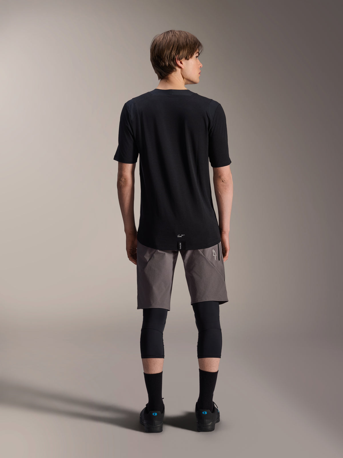 Alpinestars A-Dura Elite Shorts, ciemnoszare spodenki rowerowe MTB, widok z tyłu, zestawione z czarną koszulką z krótkim rękawem, nakolannikami i czarnymi skarpetkami, techniczna odzież rowerowa do jazdy w terenie