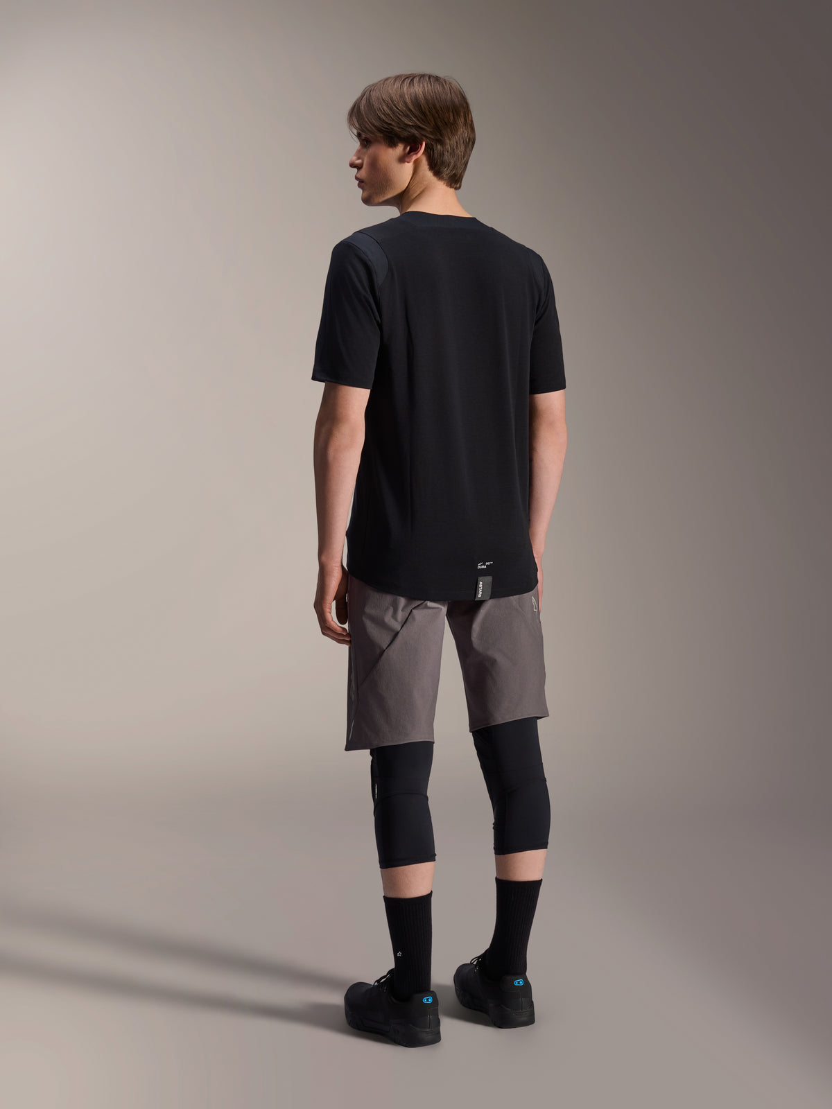 Koszulka z krótkim rękawem Alpinestars A-Dura Dri Wool i spodenki A-Dura Elite, czarna i ciemnoszara odzież do jazdy na rowerze górskim, zestaw z czarnymi ochraniaczami kolan i skarpetami, widok z tyłu na sprzęt rowerowy do jazdy w terenie