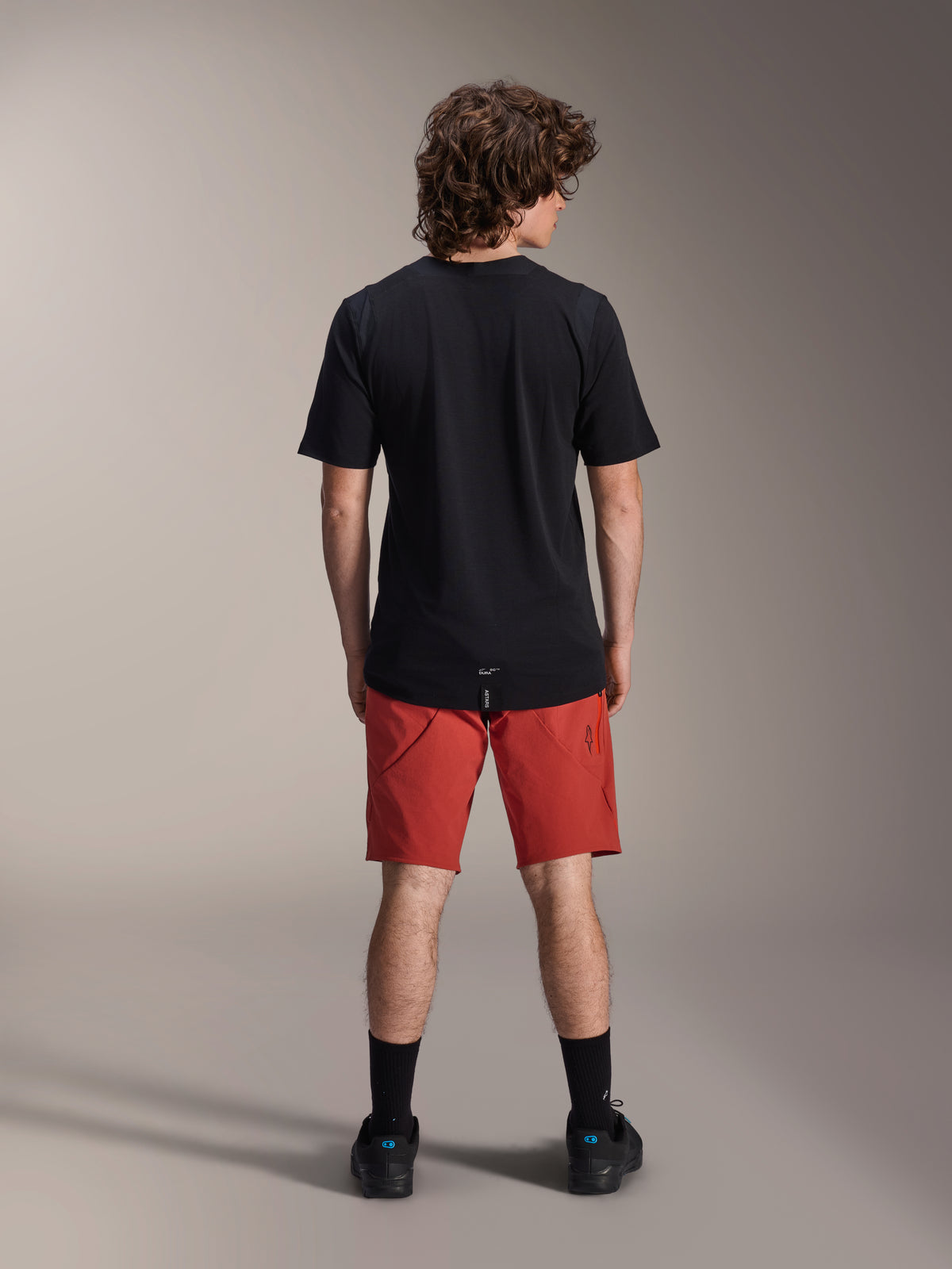 Model w spodenkach Alpinestars A-Dura Elite w kolorze Dark Orange i czarnej koszulce z krótkim rękawem A-Dura Dri Wool, widok od tyłu, w komplecie z czarnymi skarpetami i butami rowerowymi, profesjonalna odzież do kolarstwa górskiego